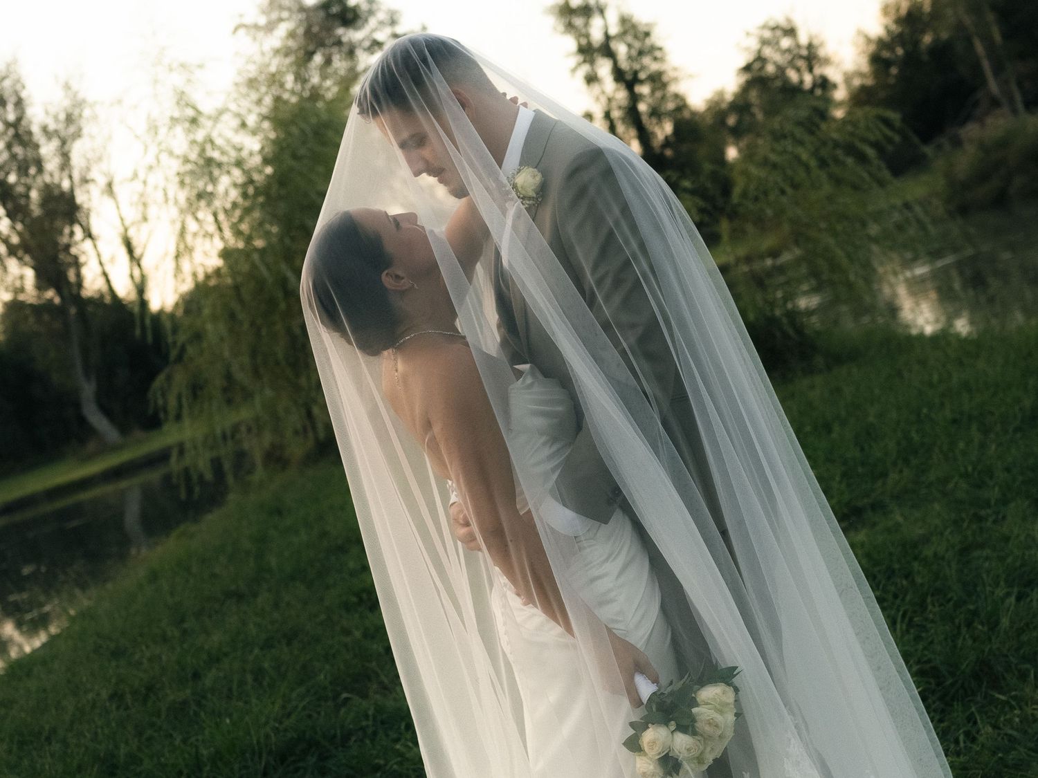 Bride and groom sharing a romantic kiss under a flowing wedding veil outdoors near a pond.