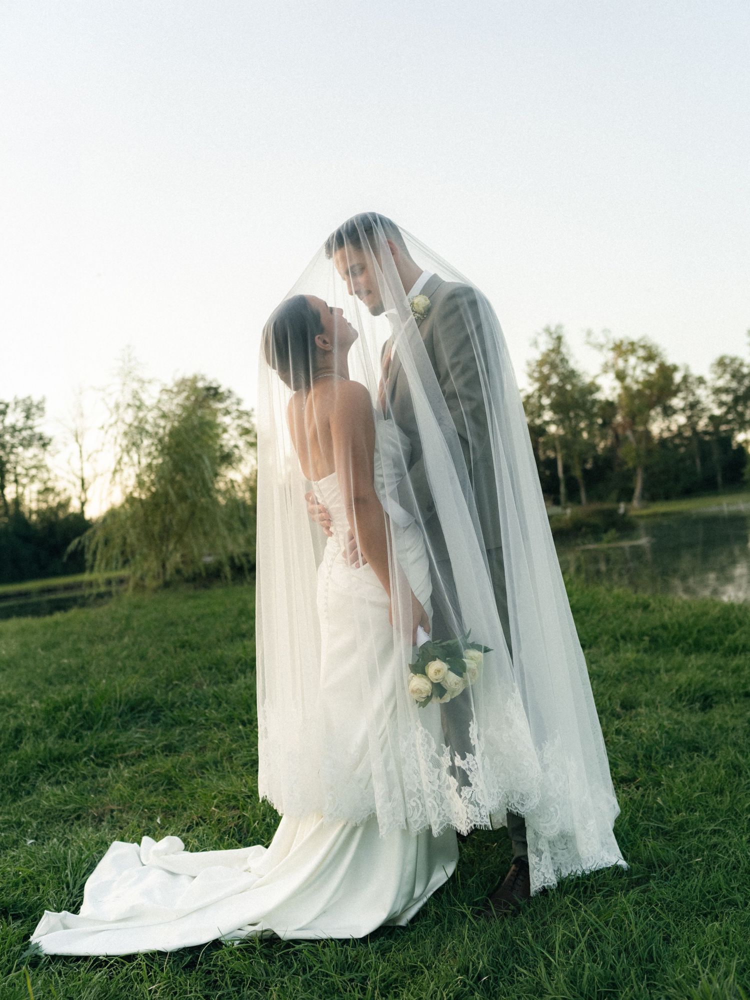 Bride and groom share a romantic moment under a flowing cathedral veil outdoors near a pond.