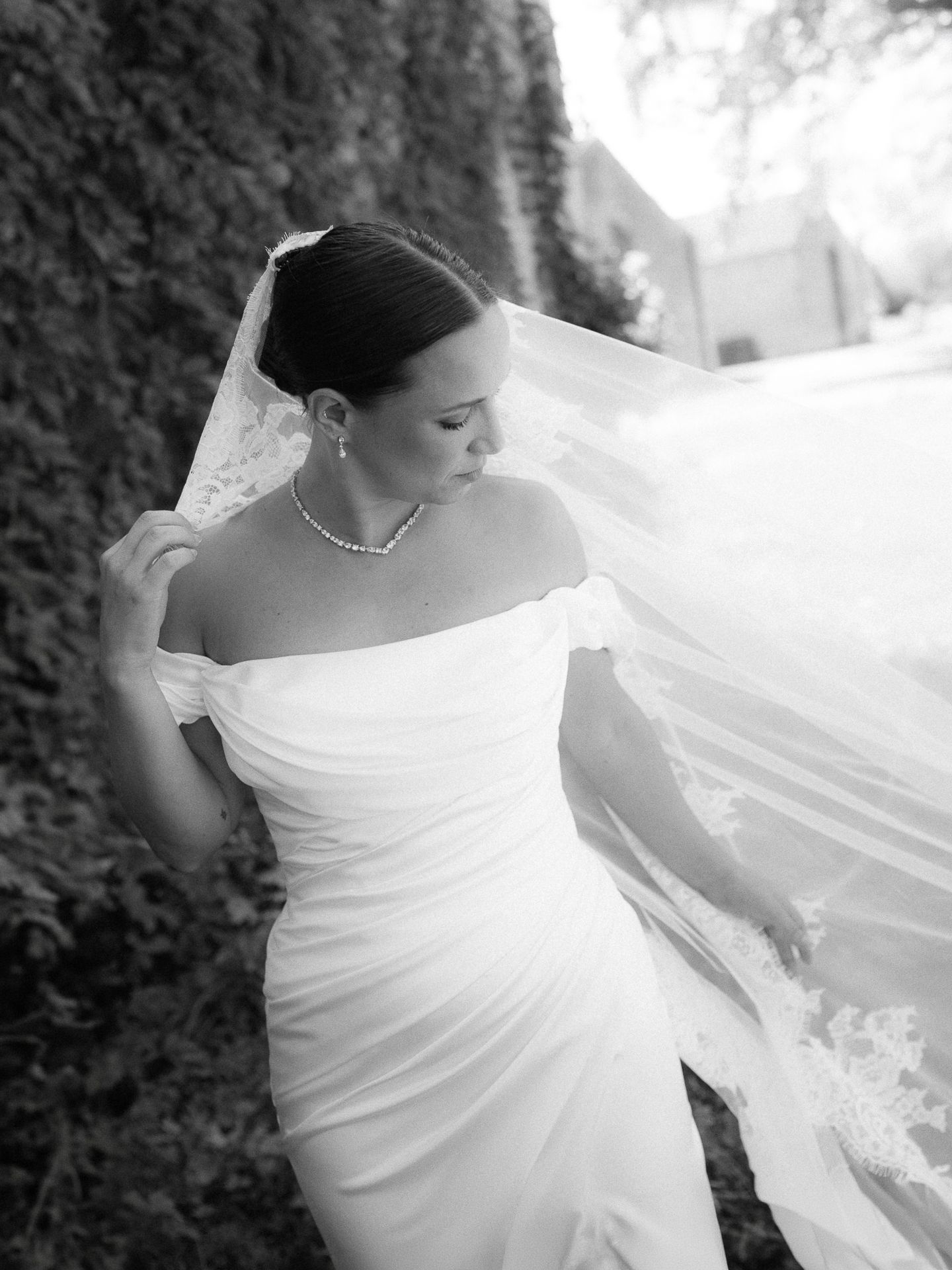 Black and white bridal portrait of a bride in an off-shoulder gown with flowing veil and pearl jewelry outdoors.