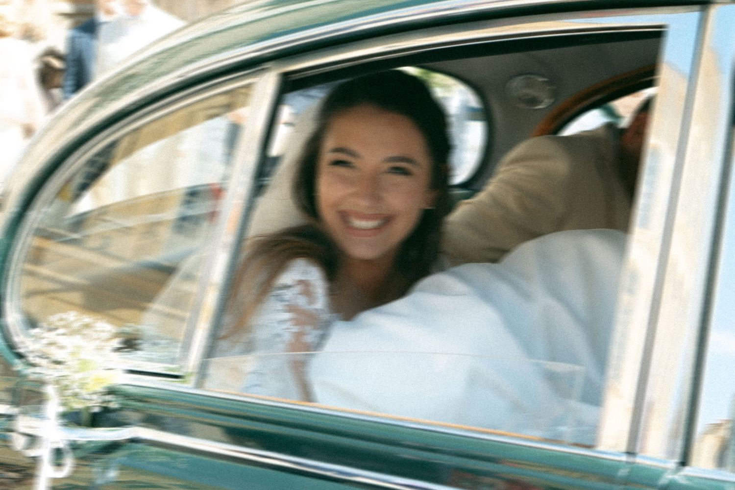 Bride wearing a white wedding dress sits in a vintage car while smiling through the window during her special day.