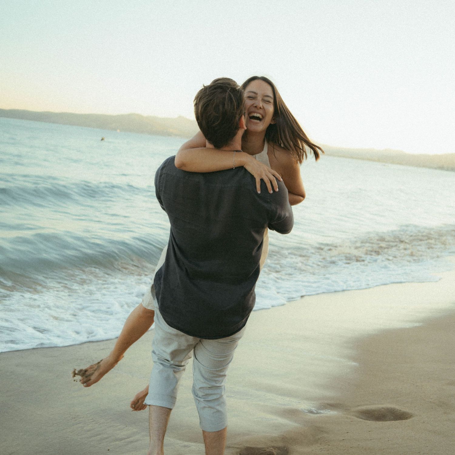 A couple embraces joyfully while playing and spinning on a sunny beach at sunset.