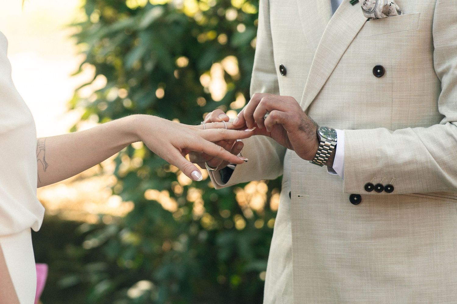 Groom in beige suit placing wedding ring on bride's finger during outdoor ceremony.