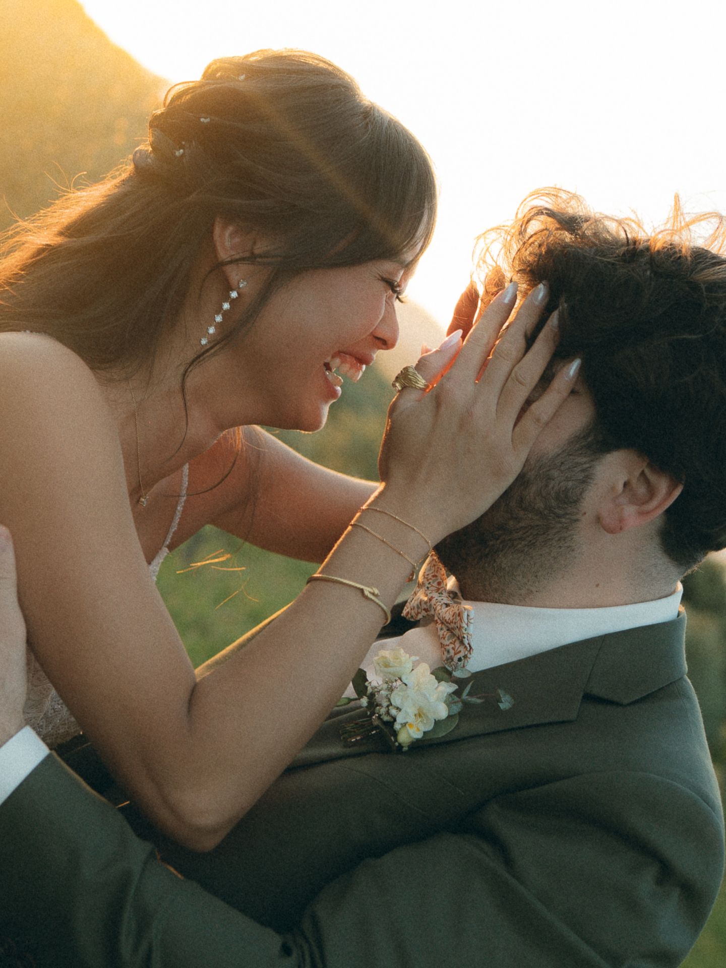 A bride and groom share a tender golden hour moment, smiling at each other as she gently holds his face outdoors.