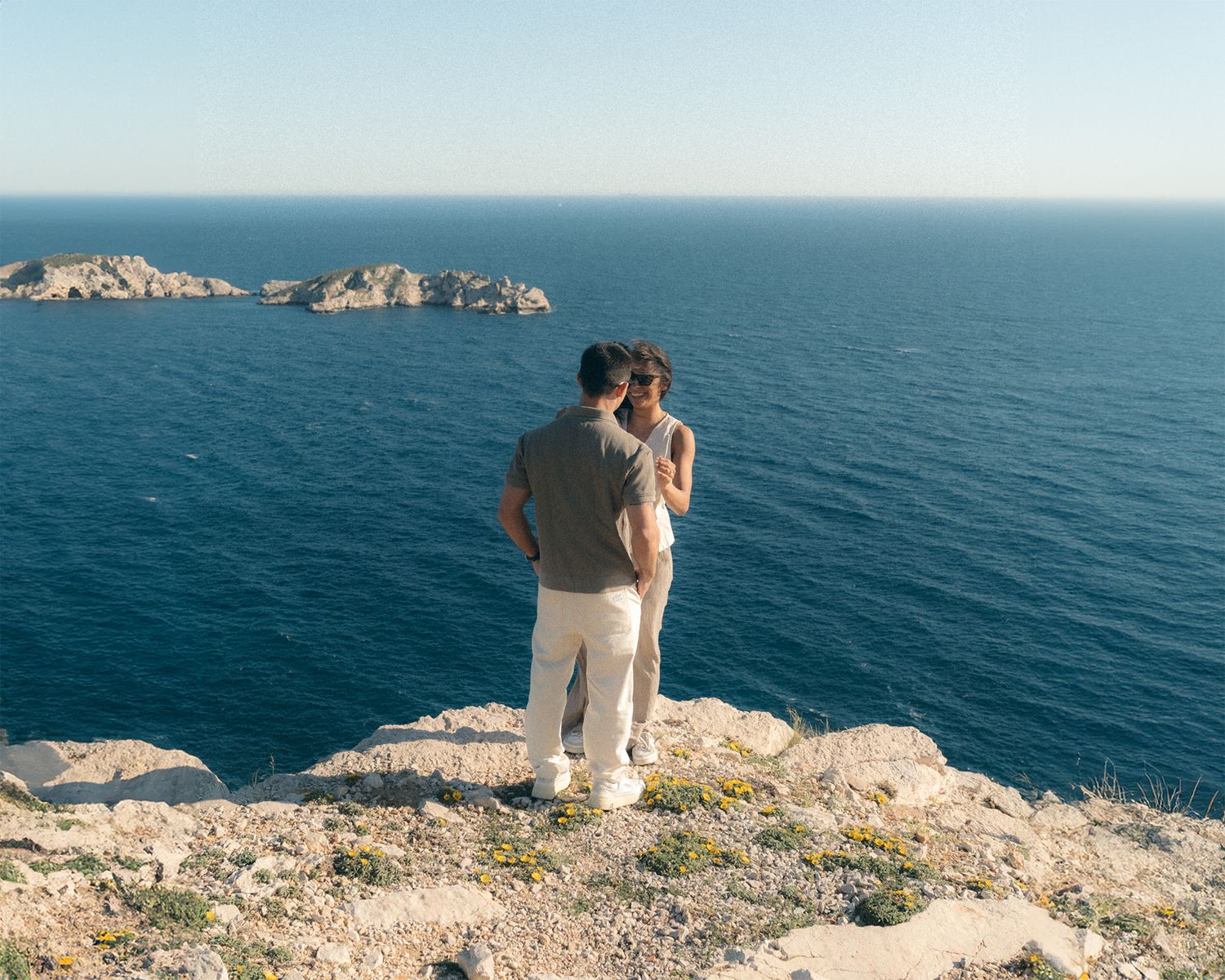 A person sits peacefully on a coastal cliff edge overlooking the vast blue Mediterranean Sea on a sunny day.
