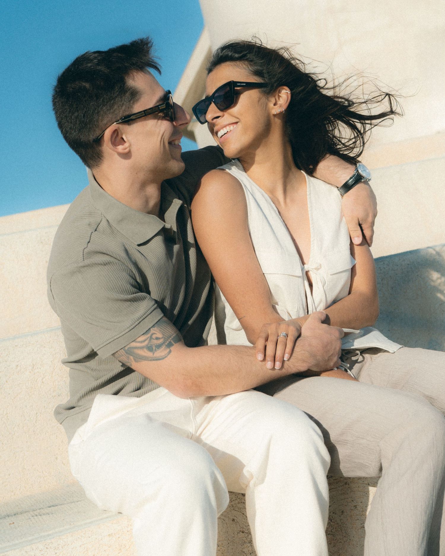 A couple in sunglasses share an intimate moment while sitting together outdoors against a bright blue sky.