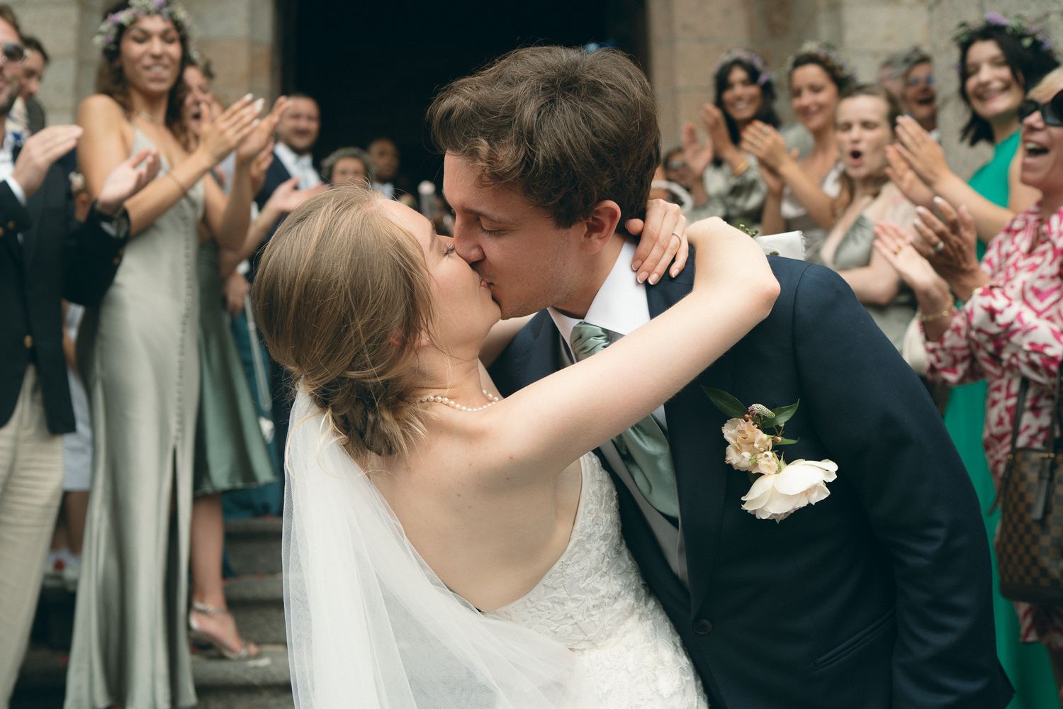 A bride and groom share a romantic kiss during their wedding celebration surrounded by joyful guests.