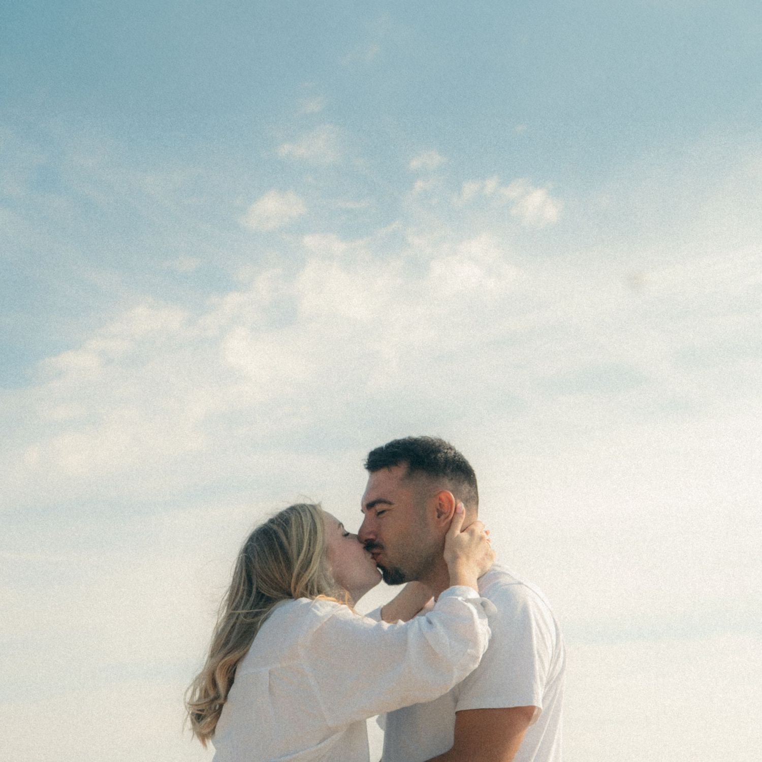 A romantic couple shares an intimate moment together against a soft cloudy sky background.