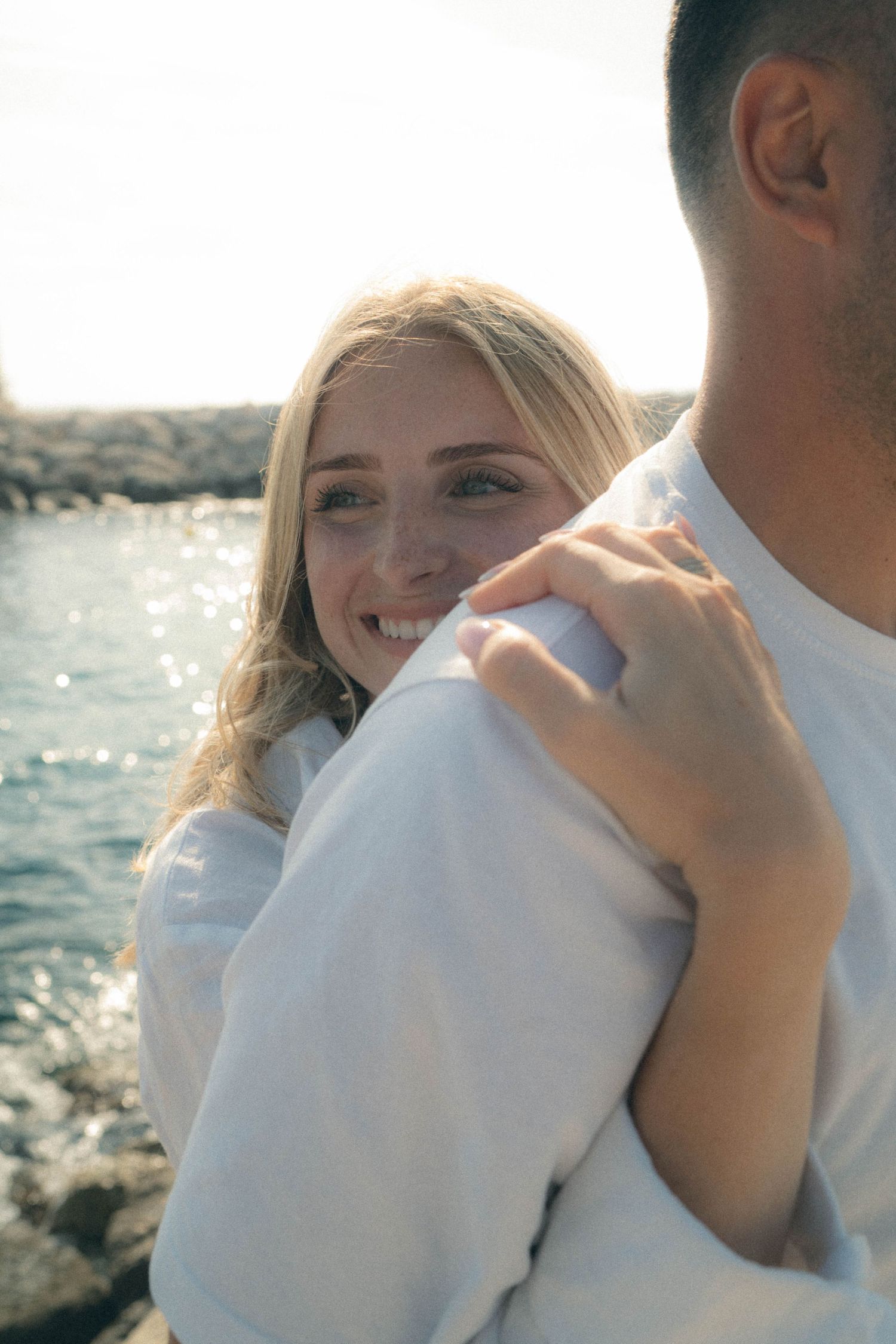 A couple embraces by the ocean with sparkling water in the background during a romantic seaside moment.