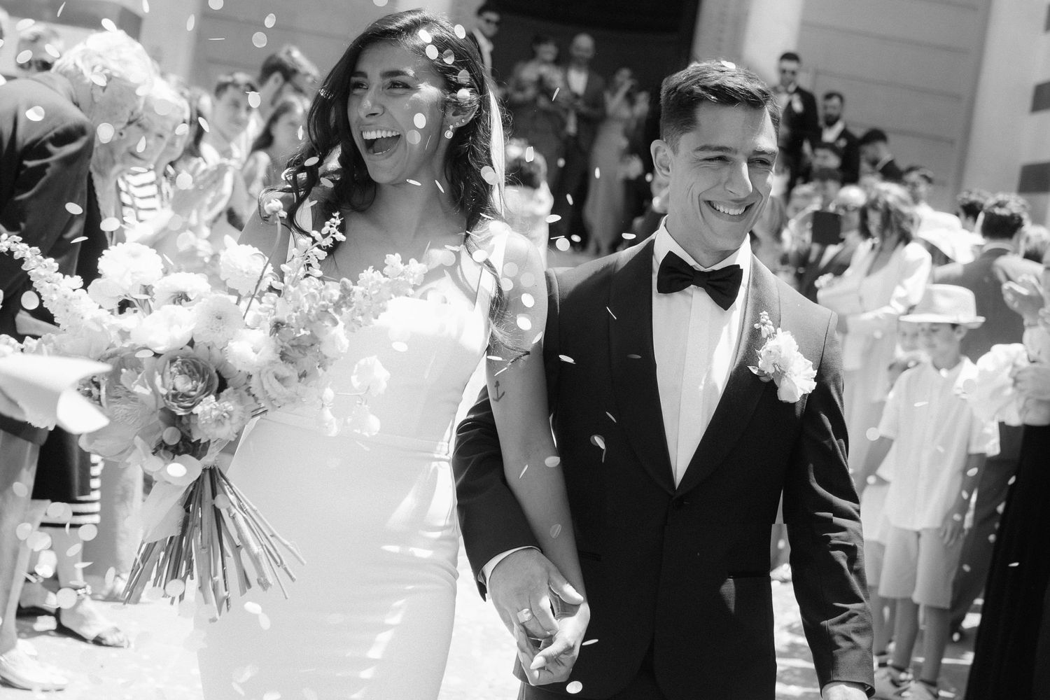 Wedding guests throw white flower petals in celebration as a couple exits their ceremony.