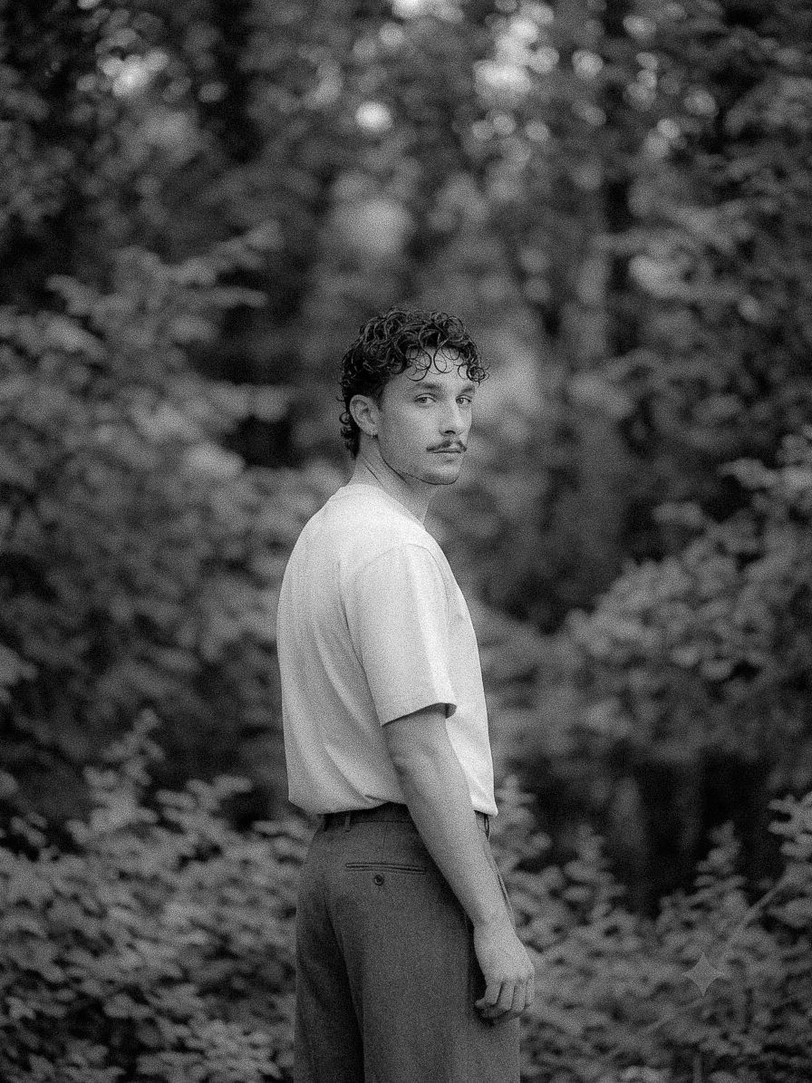 Black and white portrait of a young man with curly hair and mustache standing in a lush forest setting.
