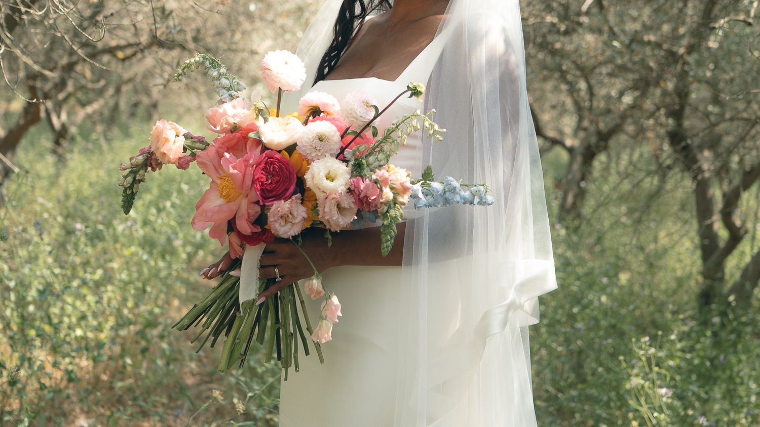 A wedding bouquet of pink and peach roses with delicate greenery is held against a flowing white dress outdoors.