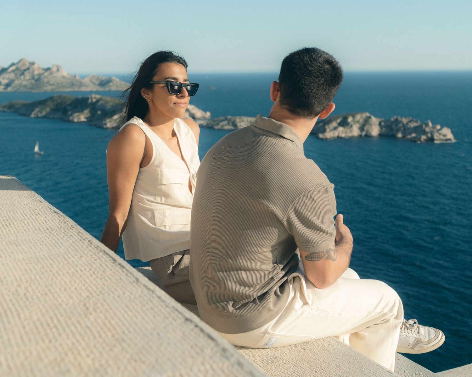 A couple sits together on a coastal cliff overlooking the blue ocean and rocky coastline on a sunny day.
