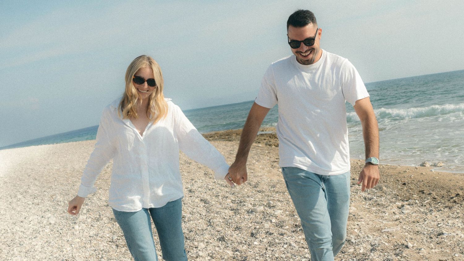 A couple in matching white shirts and jeans walks hand in hand along a sunny beach shoreline.