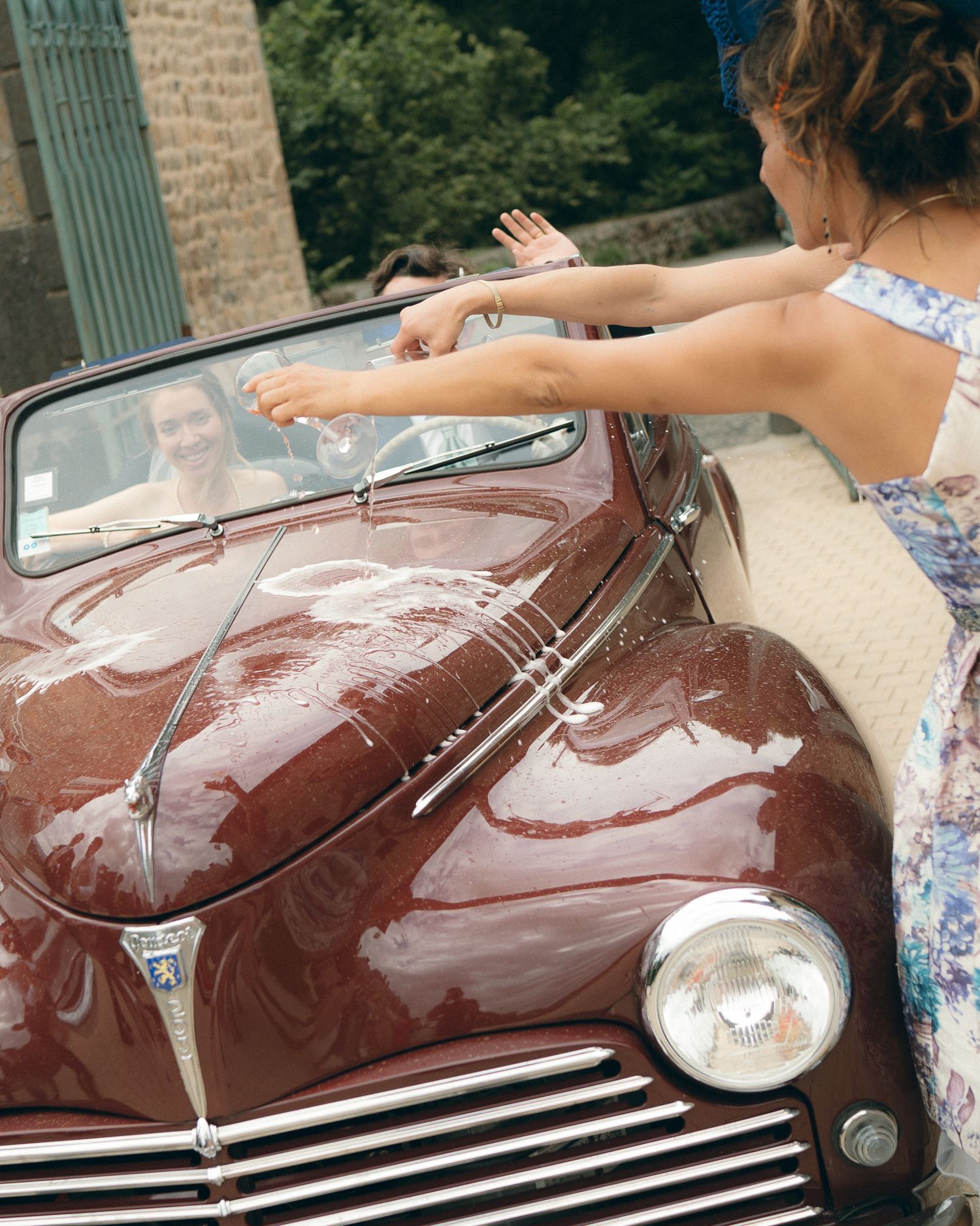A person washes a vintage burgundy car by hand using soapy water and a cloth in a driveway.