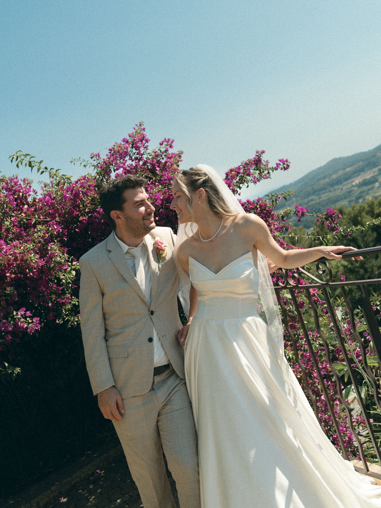 Bride and groom share a romantic moment on a balcony surrounded by vibrant pink bougainvillea with mountain views.