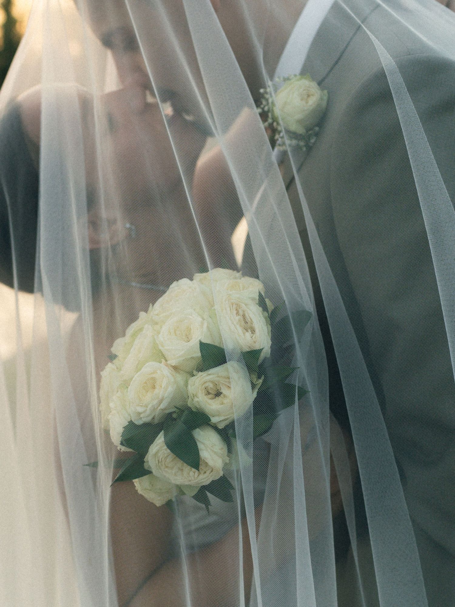 Bride and groom share an intimate moment under a flowing white veil, bride holding a bouquet of white roses.
