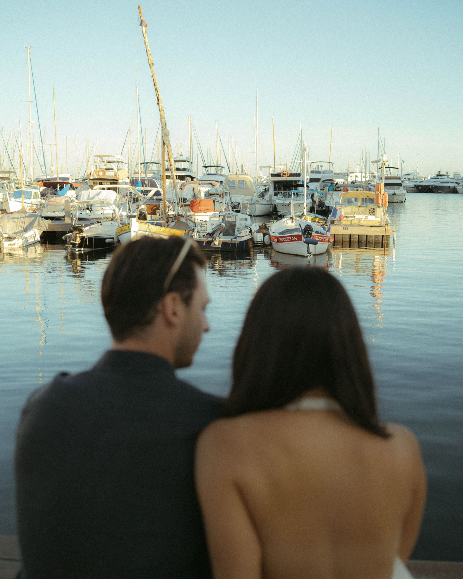 A couple stands silhouetted against a marina backdrop with boats and masts visible in the calm water at sunset.