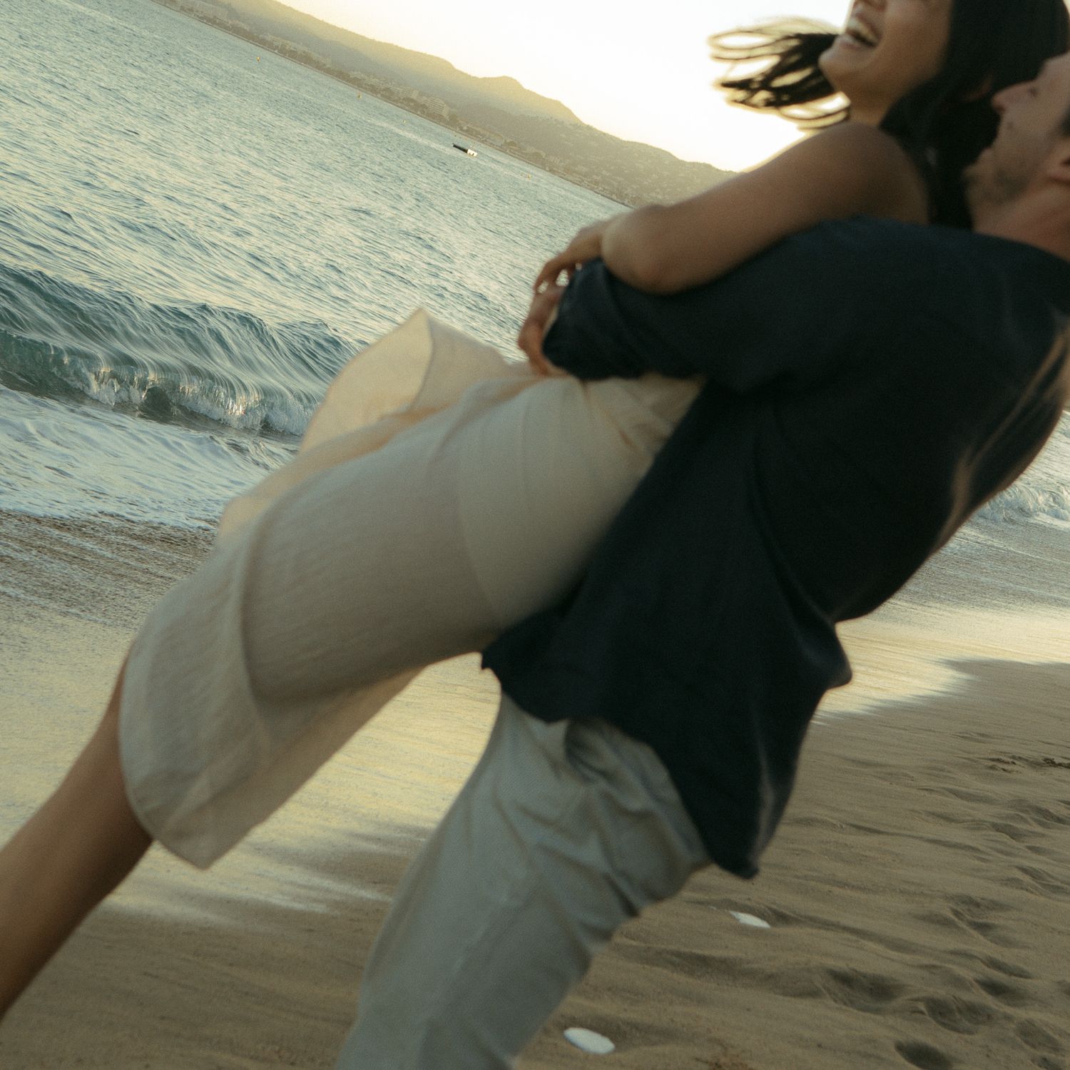 Couple sharing an intimate dance moment on a sandy beach at sunset with ocean waves in the background.