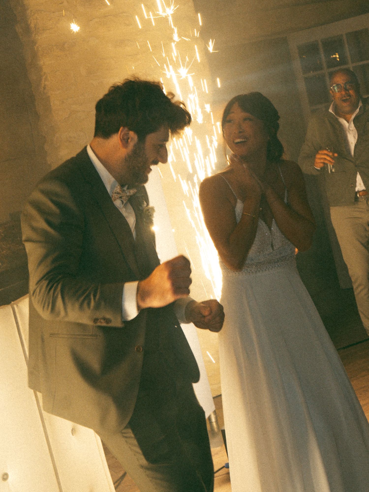 Bride and groom dancing joyfully at wedding reception with sparkling fireworks in the background.