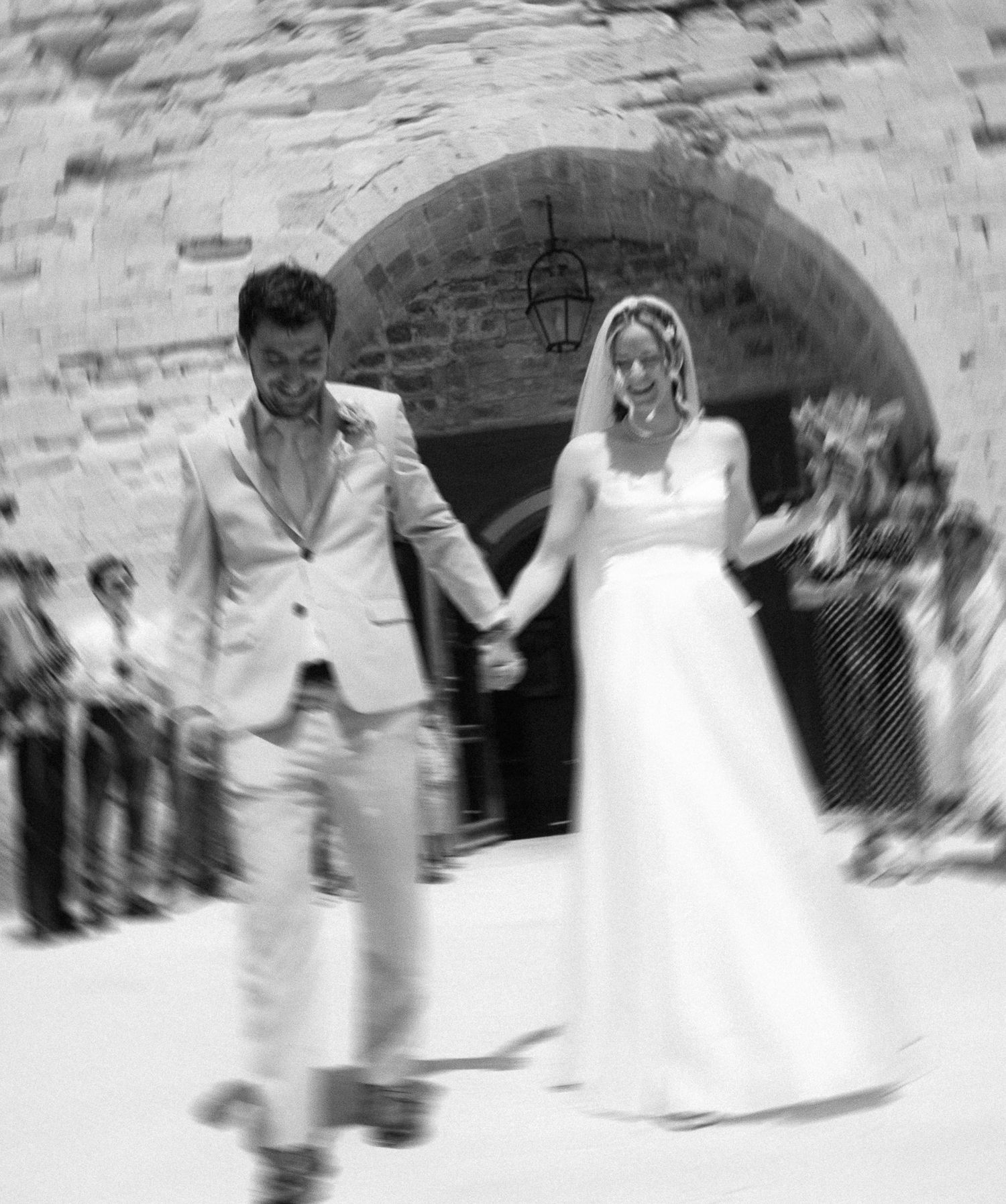 Black and white photo of a bride and groom holding hands, walking out of a stone arch church after their wedding ceremony.
