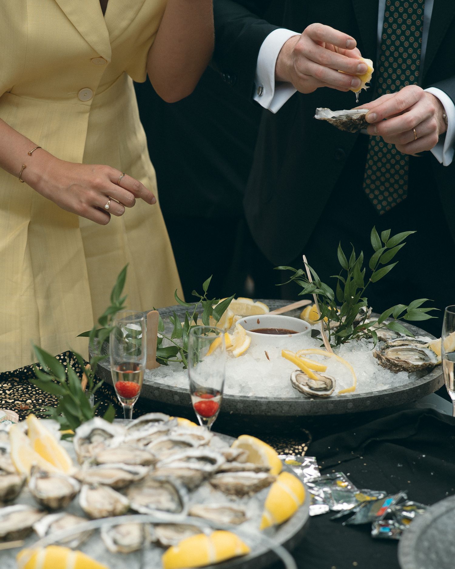 A seafood platter with fresh oysters and lemon slices is displayed on a dark table with hands serving in formal attire.