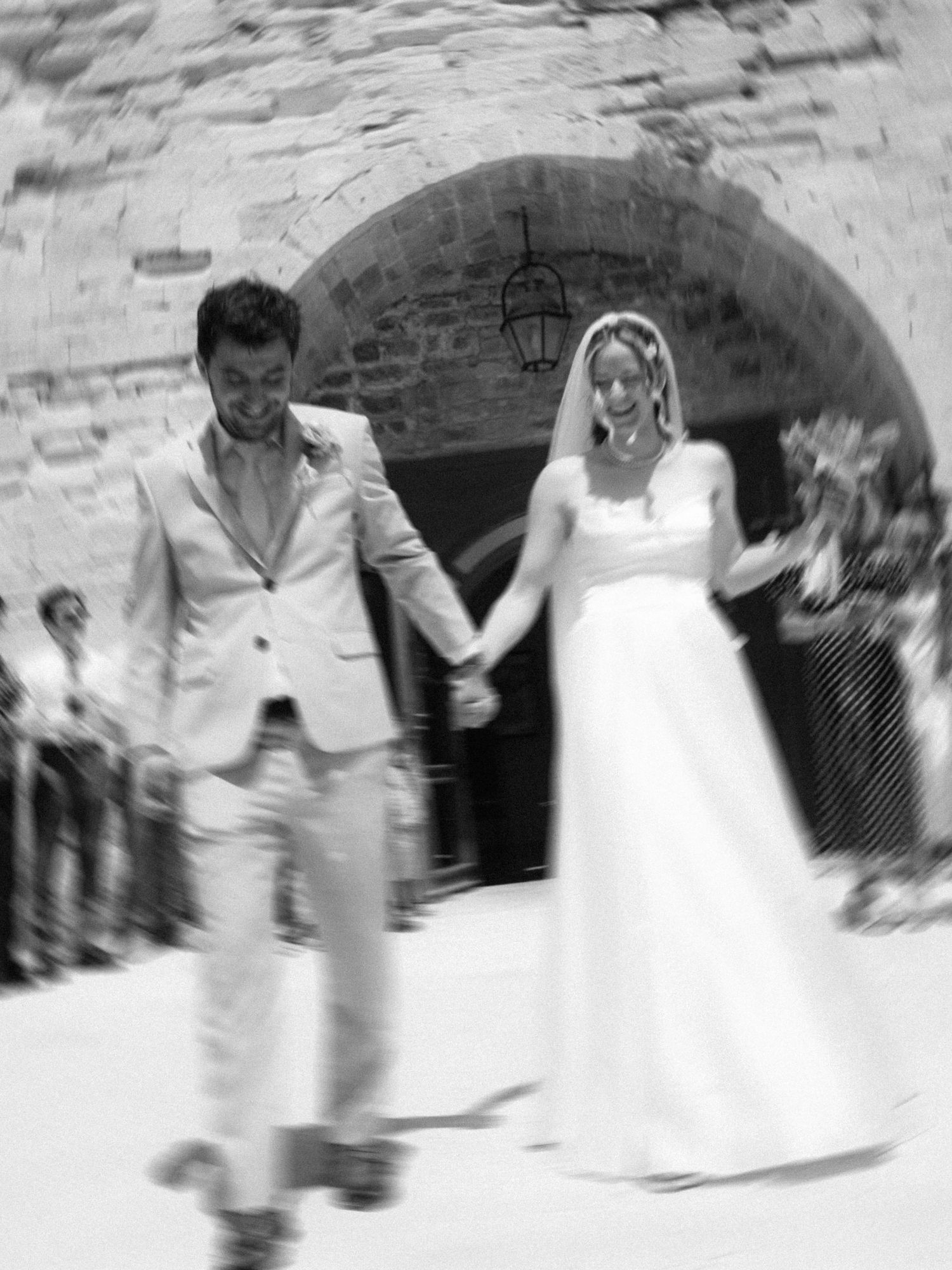 Black and white photo of a bride and groom holding hands, walking through a stone archway at their wedding.