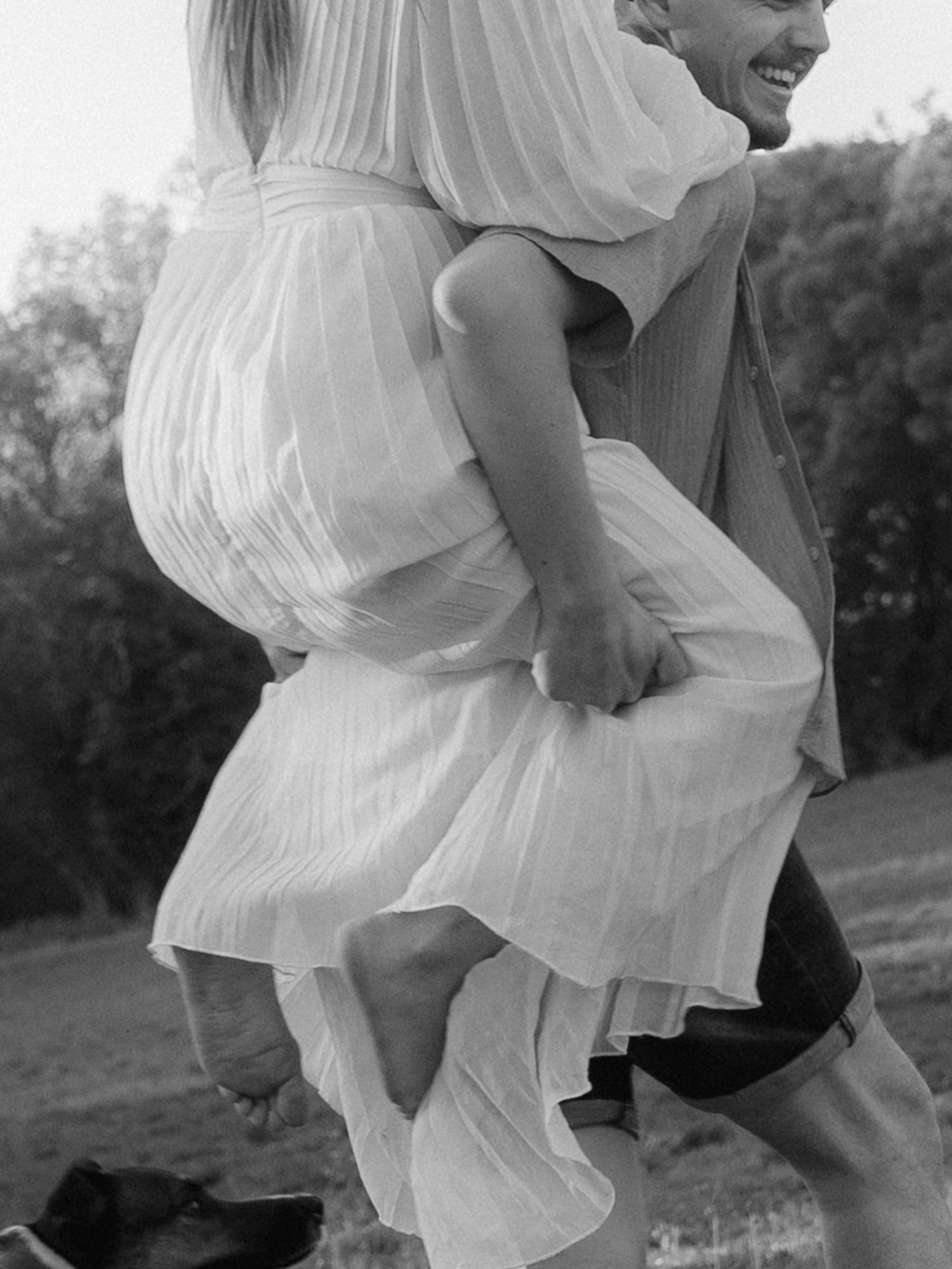 Black and white photo of a laughing man carrying a woman in a white dress on his back outdoors.