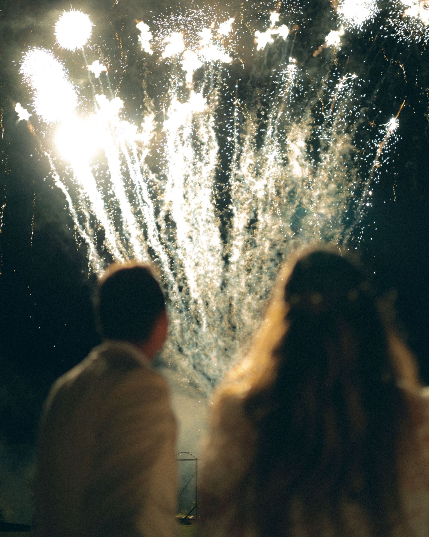 Silhouettes of people watching bright white fireworks exploding against dark night sky.