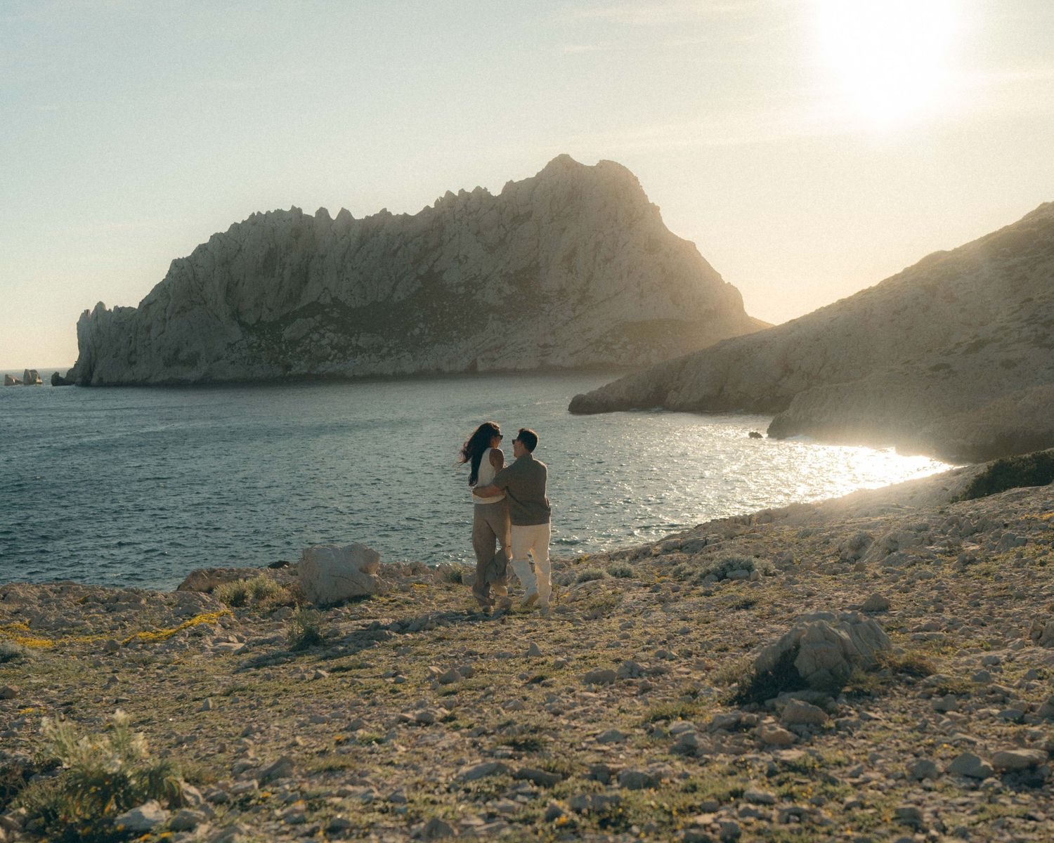 Two silhouettes stand together on a rocky Mediterranean coastline at sunset with mountains and sea in the background.