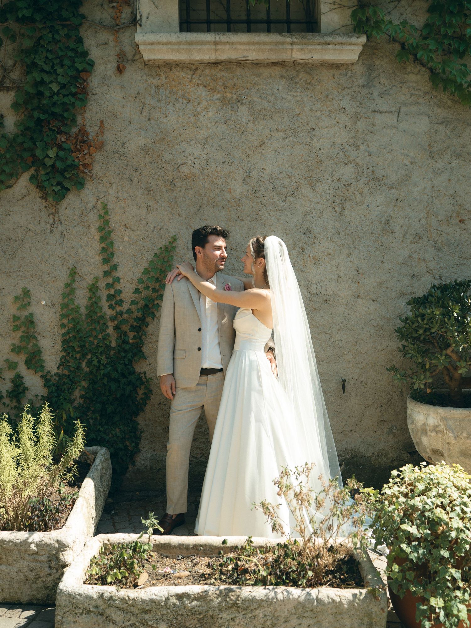 A bride and groom share a romantic moment outside a rustic stone villa surrounded by lush greenery on their wedding day.