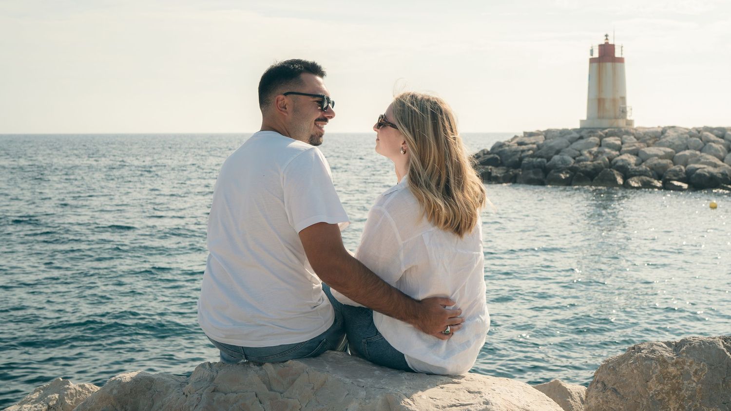 Couple sitting on rocky shoreline overlooking lighthouse at sunset.