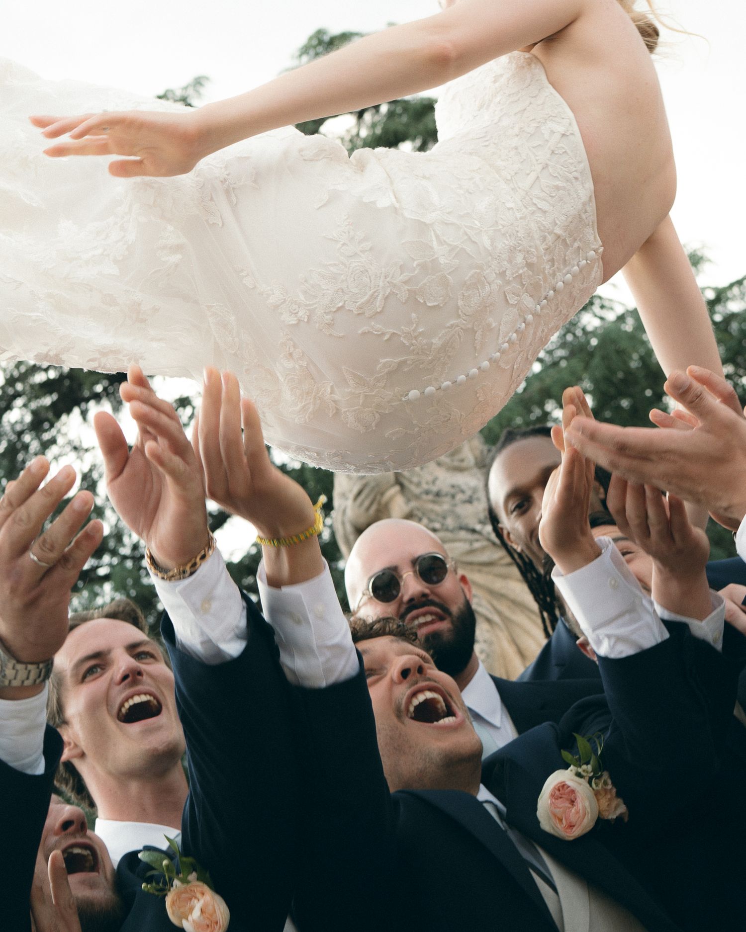 Wedding guests lift and toss a person wearing a white dress in celebration.