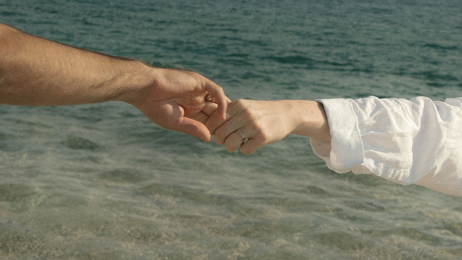A hand holds a white paper boat over rippling blue ocean water on a sunny day.