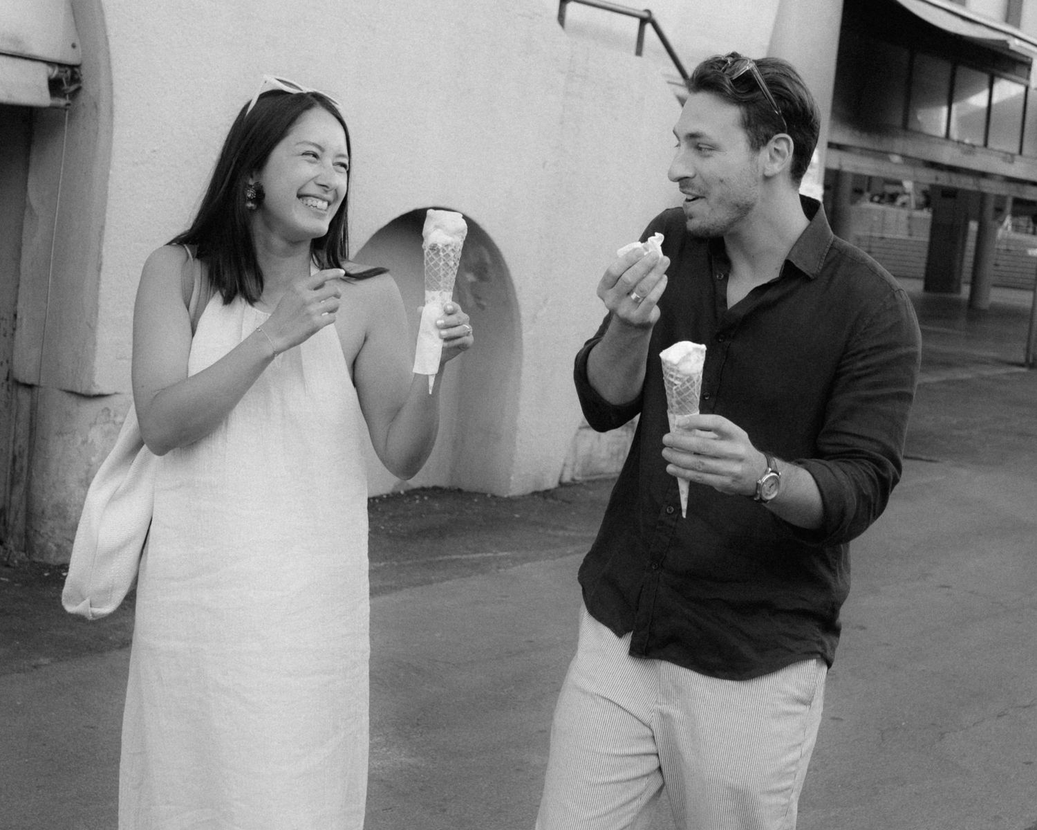 A couple shares a lighthearted moment while eating ice cream cones outdoors in black and white photography.