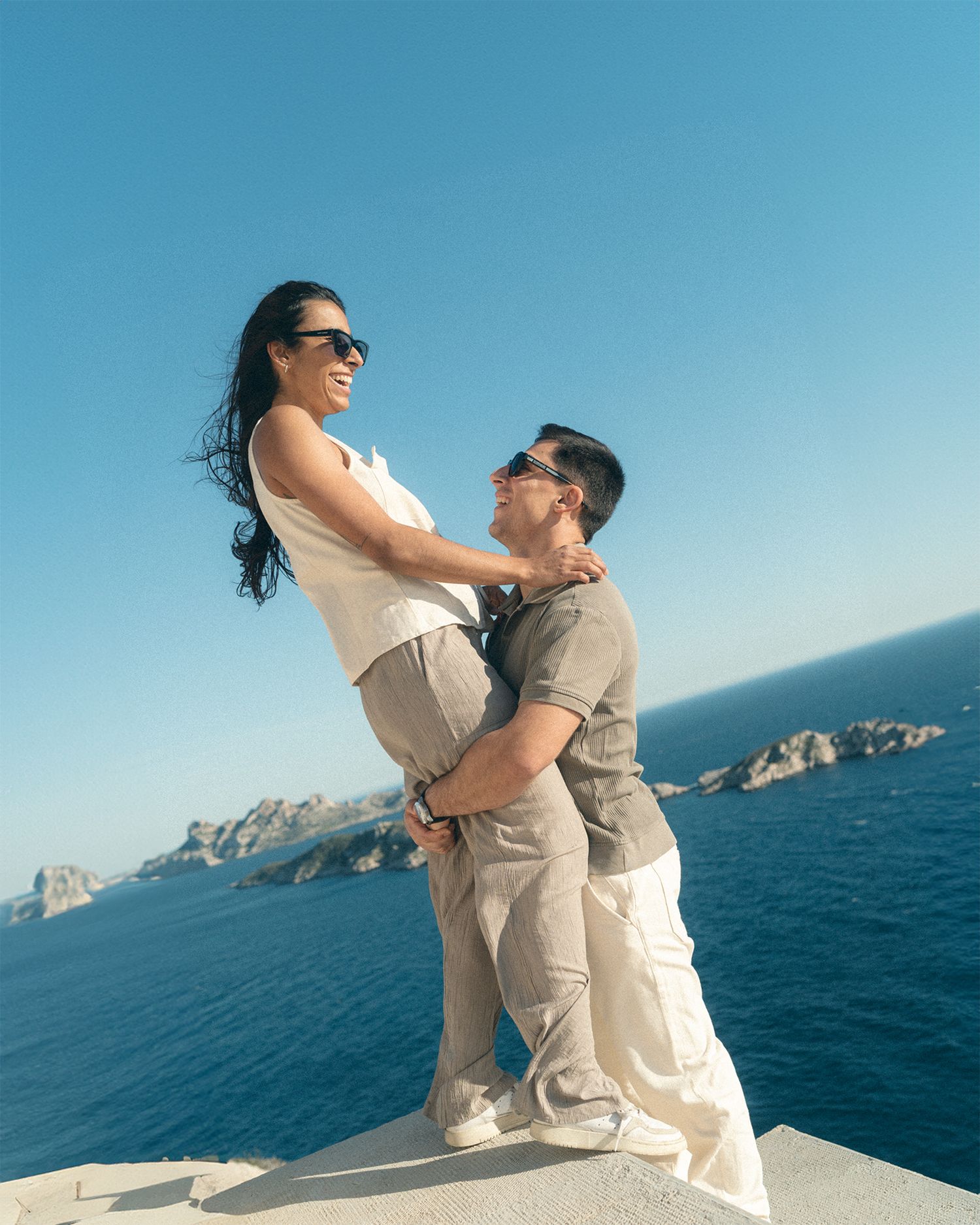 A couple embraces on a scenic cliff overlooking the ocean and rocky coastline on a bright sunny day.