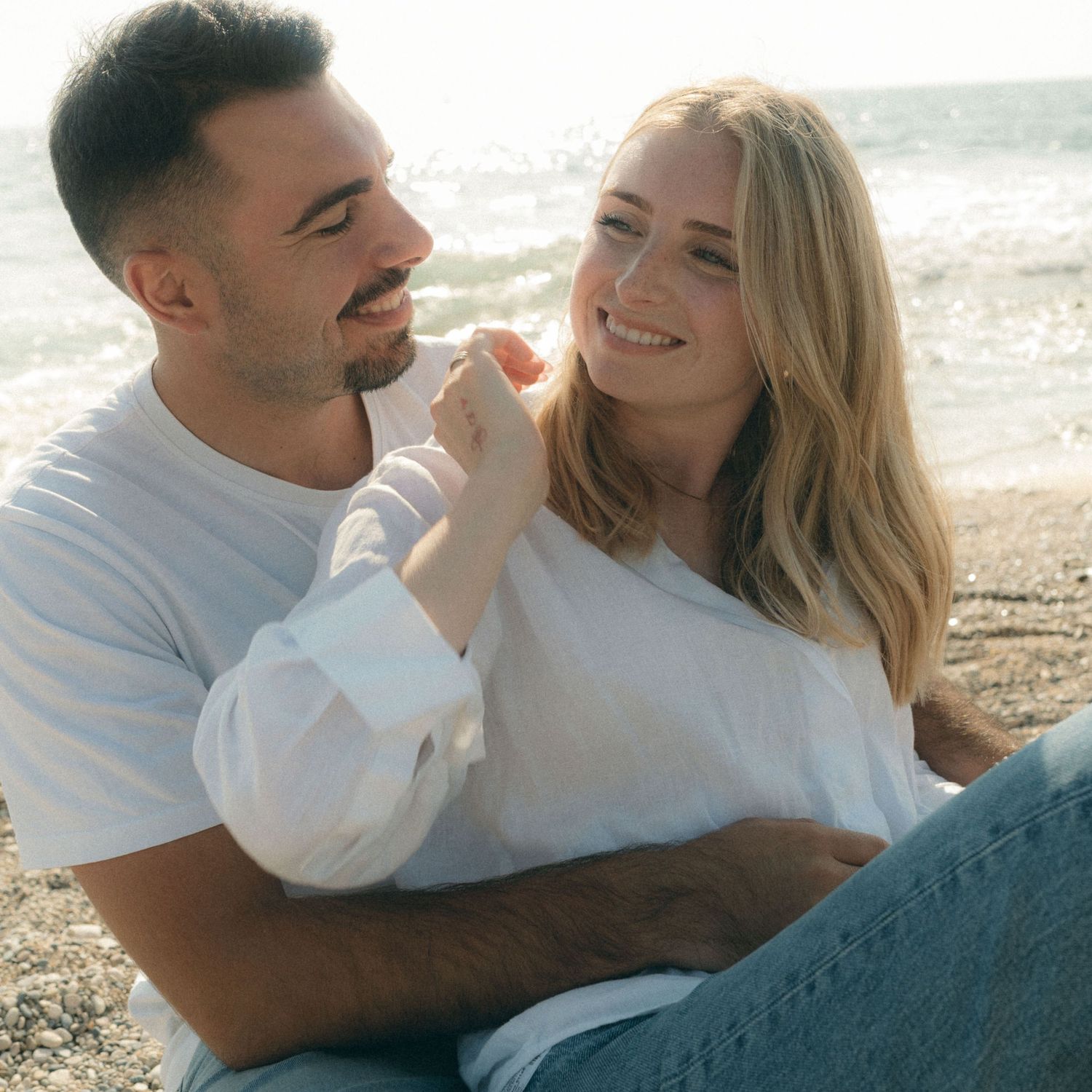A couple shares joyful moments together lying on a beach during sunset, wearing casual white clothing.