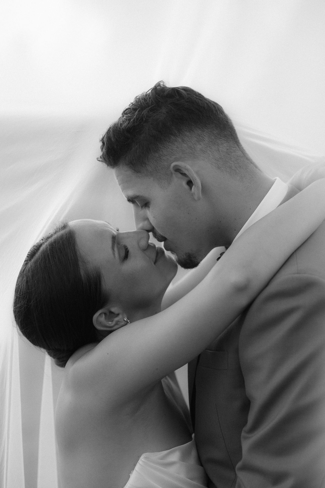 Black and white photo of a bride and groom sharing a romantic kiss under a flowing veil on their wedding day.