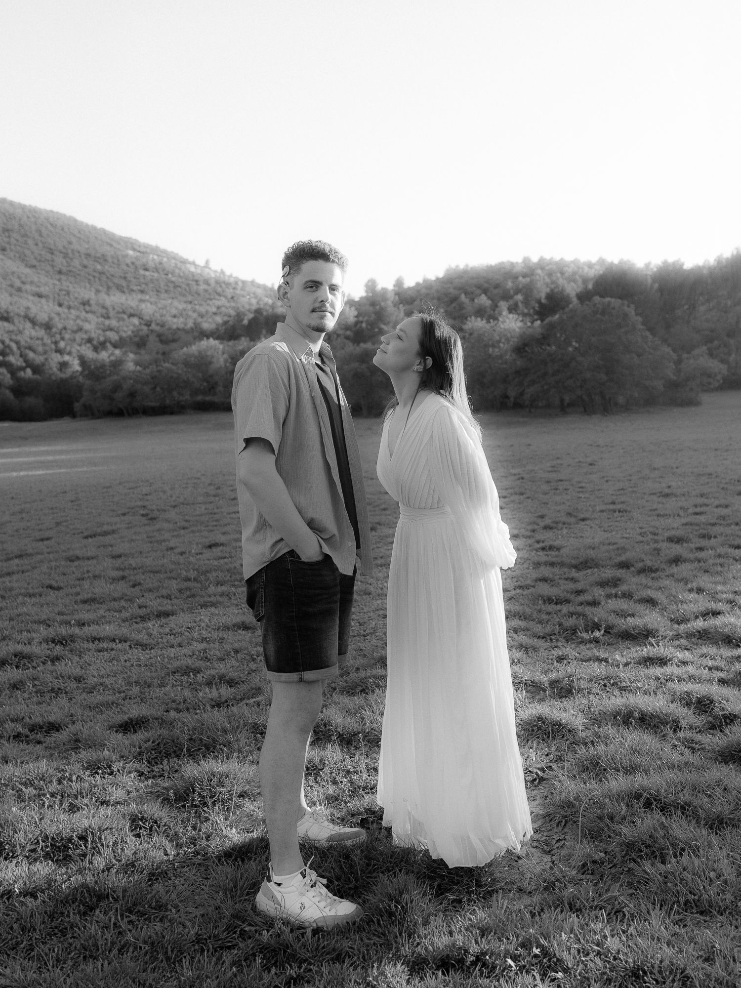 Black and white photo of a couple standing in a field, woman in white dress looking up at tall man, hills behind.