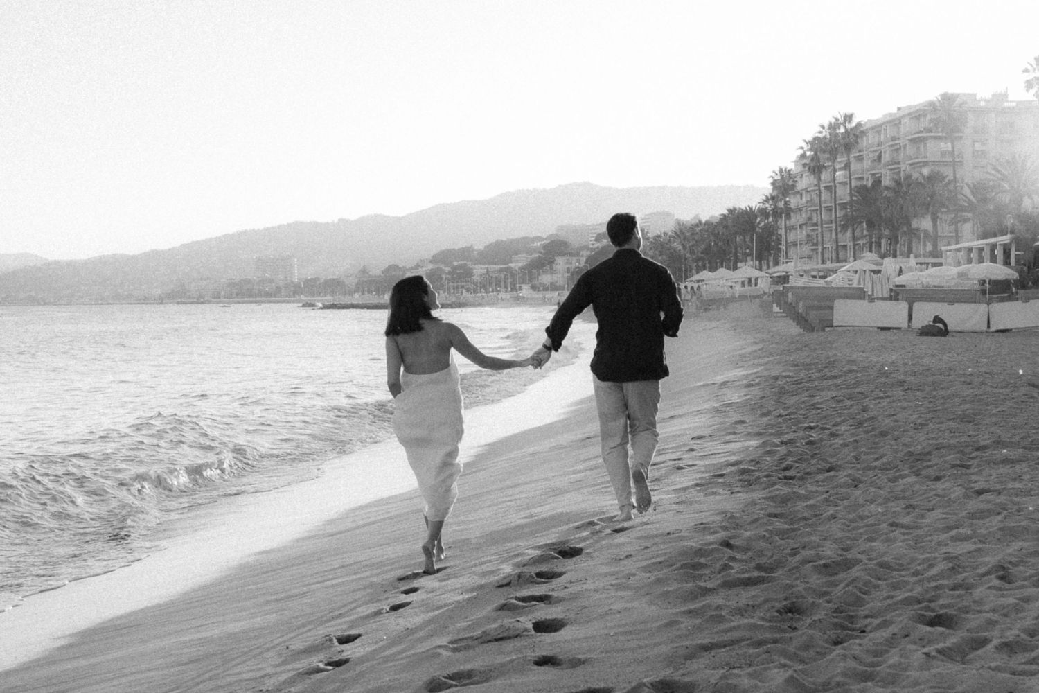 A couple holding hands while walking along a beach shoreline leaving footprints in black and white photography.