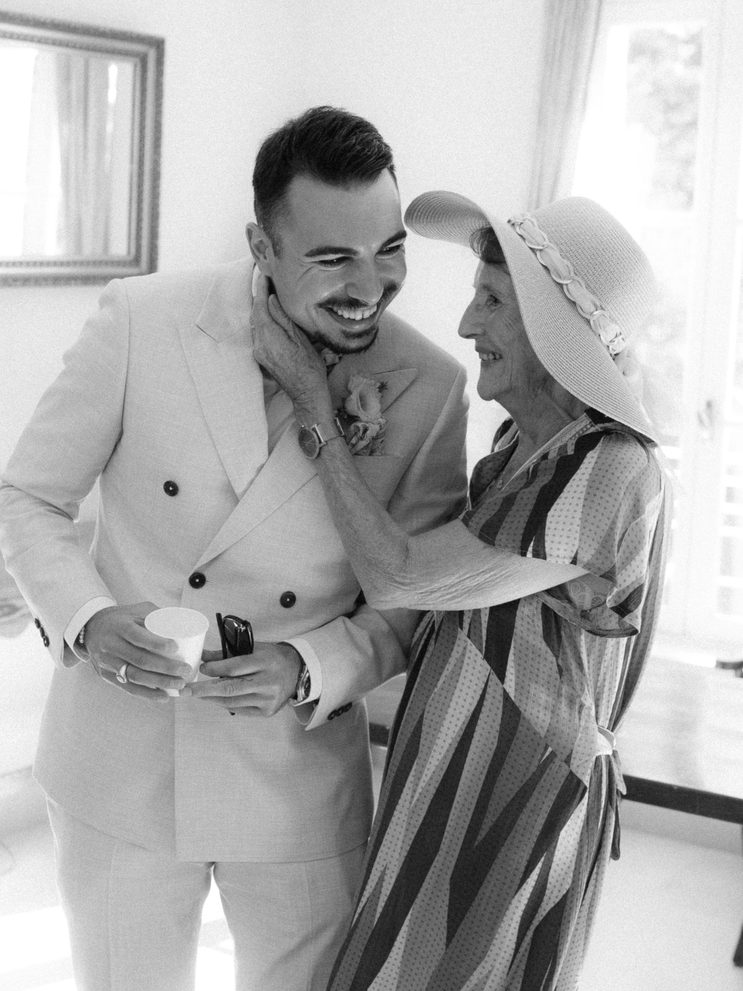 Black and white photo of a groom in a suit laughing with an older woman in a striped dress and sun hat.