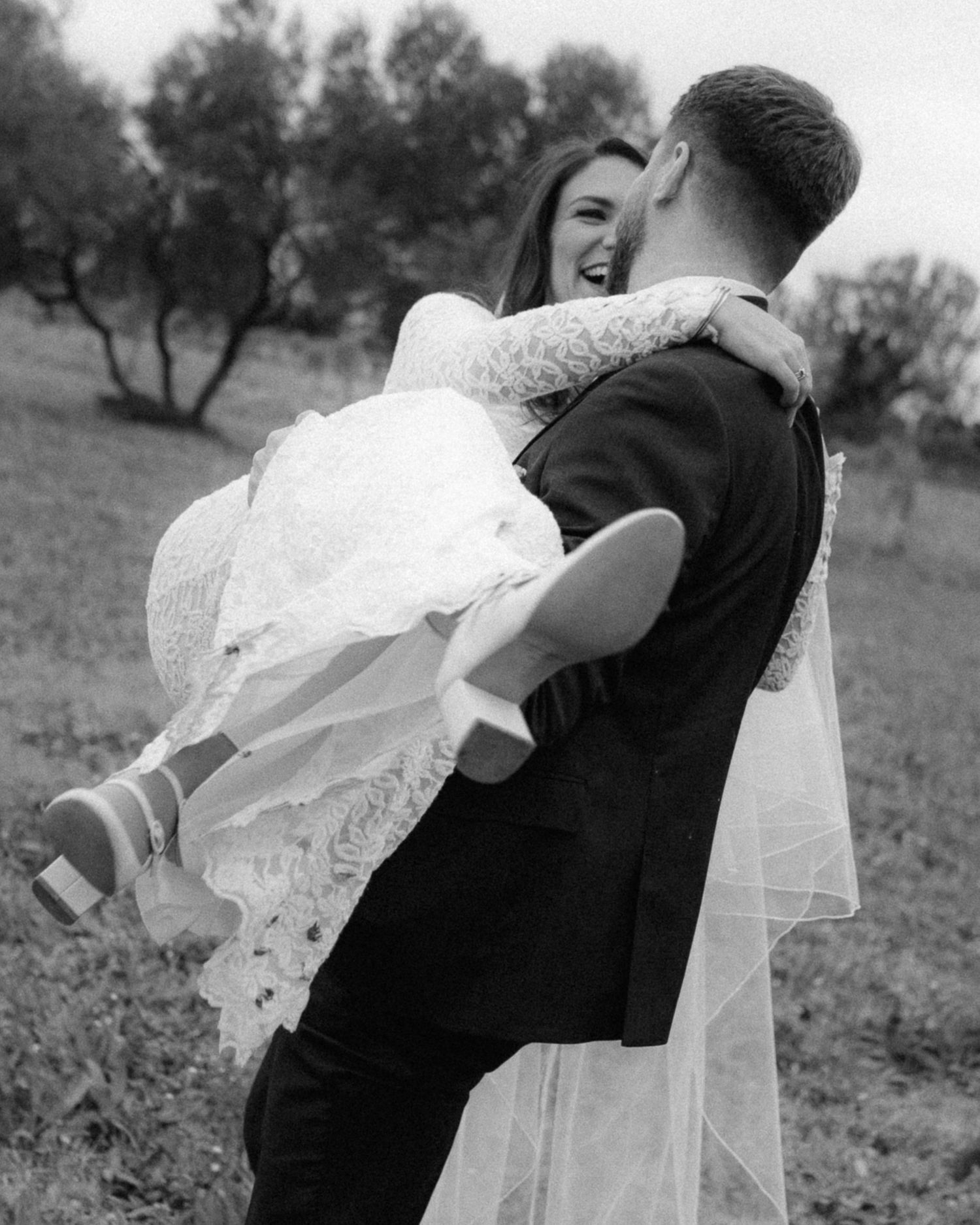 A joyful black and white wedding photo showing a couple embracing outdoors with the bride in a lace dress and flowing veil.
