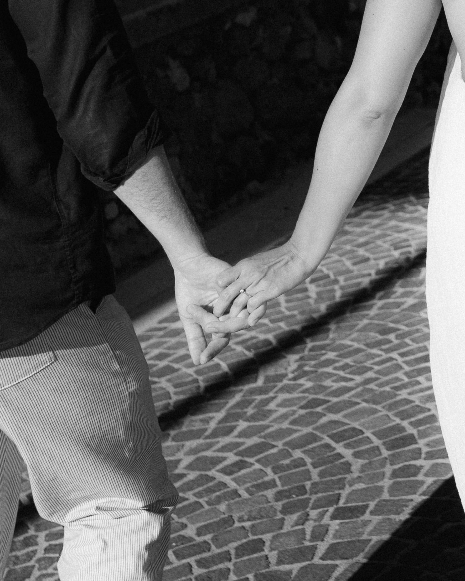 Black and white image of two hands holding each other tenderly over a cobblestone street.