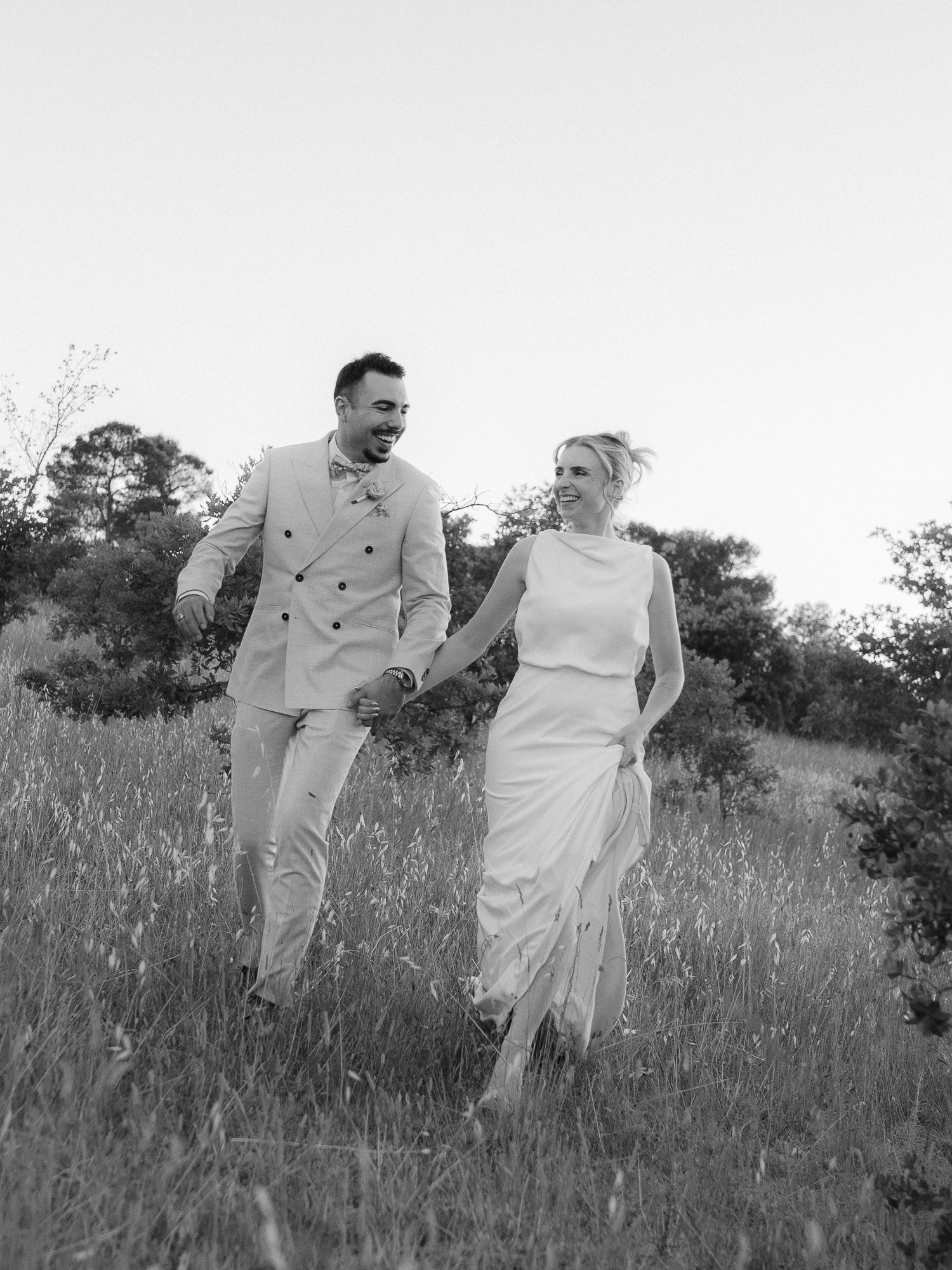 Black and white photo of a smiling couple holding hands walking through a meadow in wedding attire.