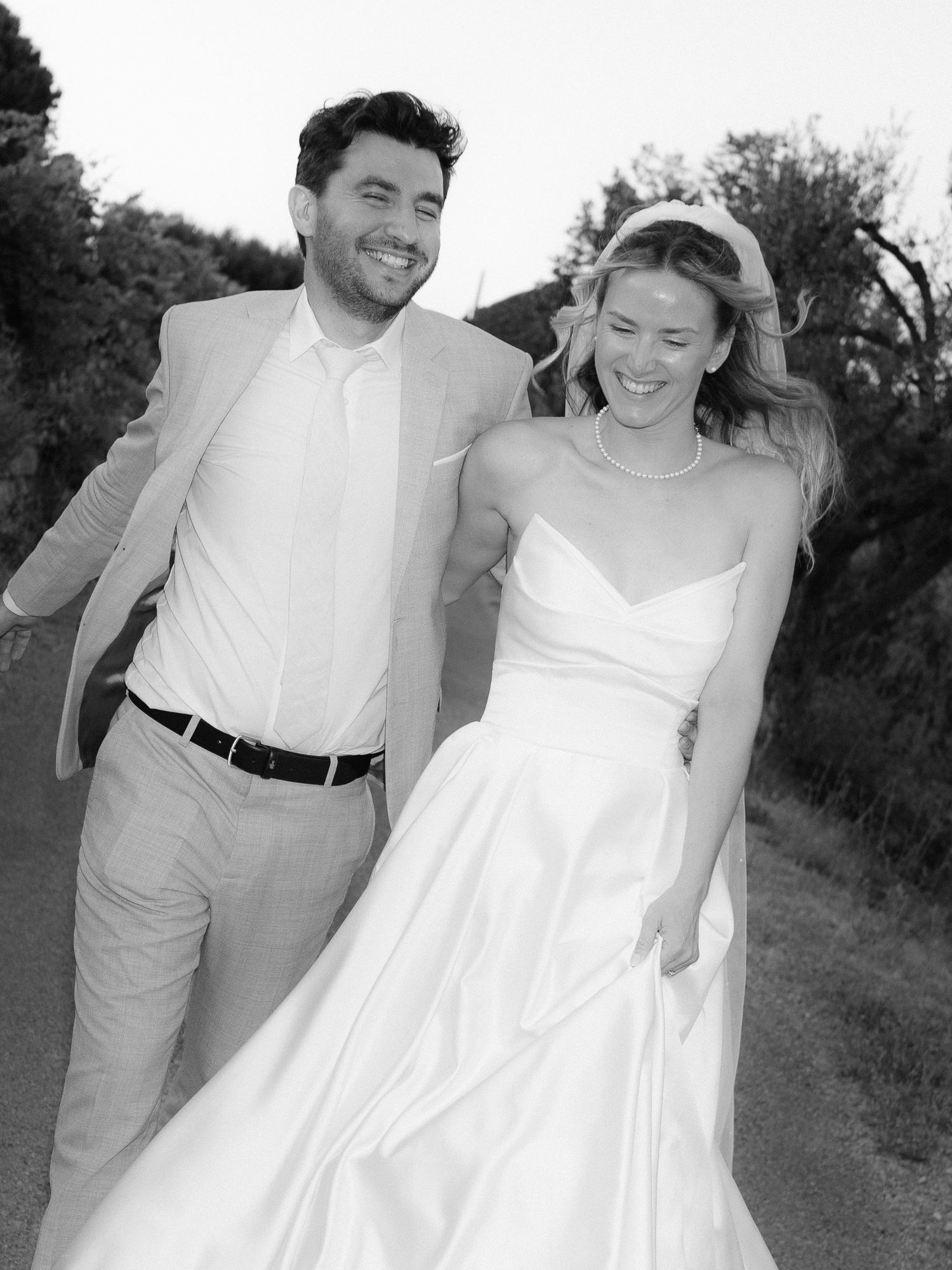 Black and white photo of a smiling bride in a white gown and groom in a suit walking together outdoors.