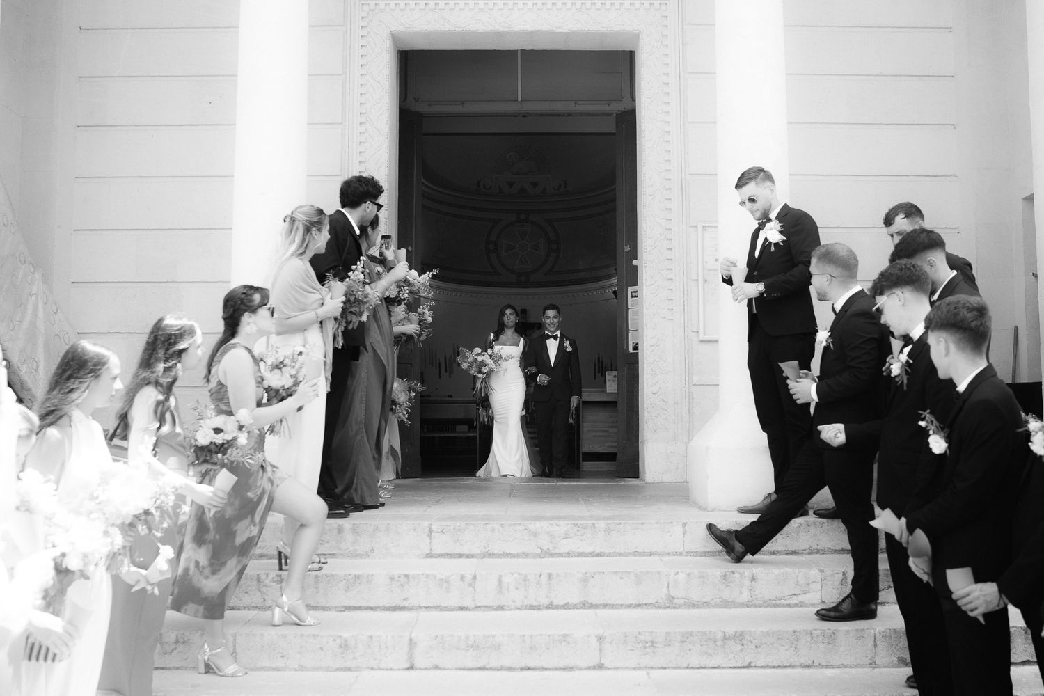 Black and white wedding photo of bride and groom exiting a grand building, greeted by guests on steps.