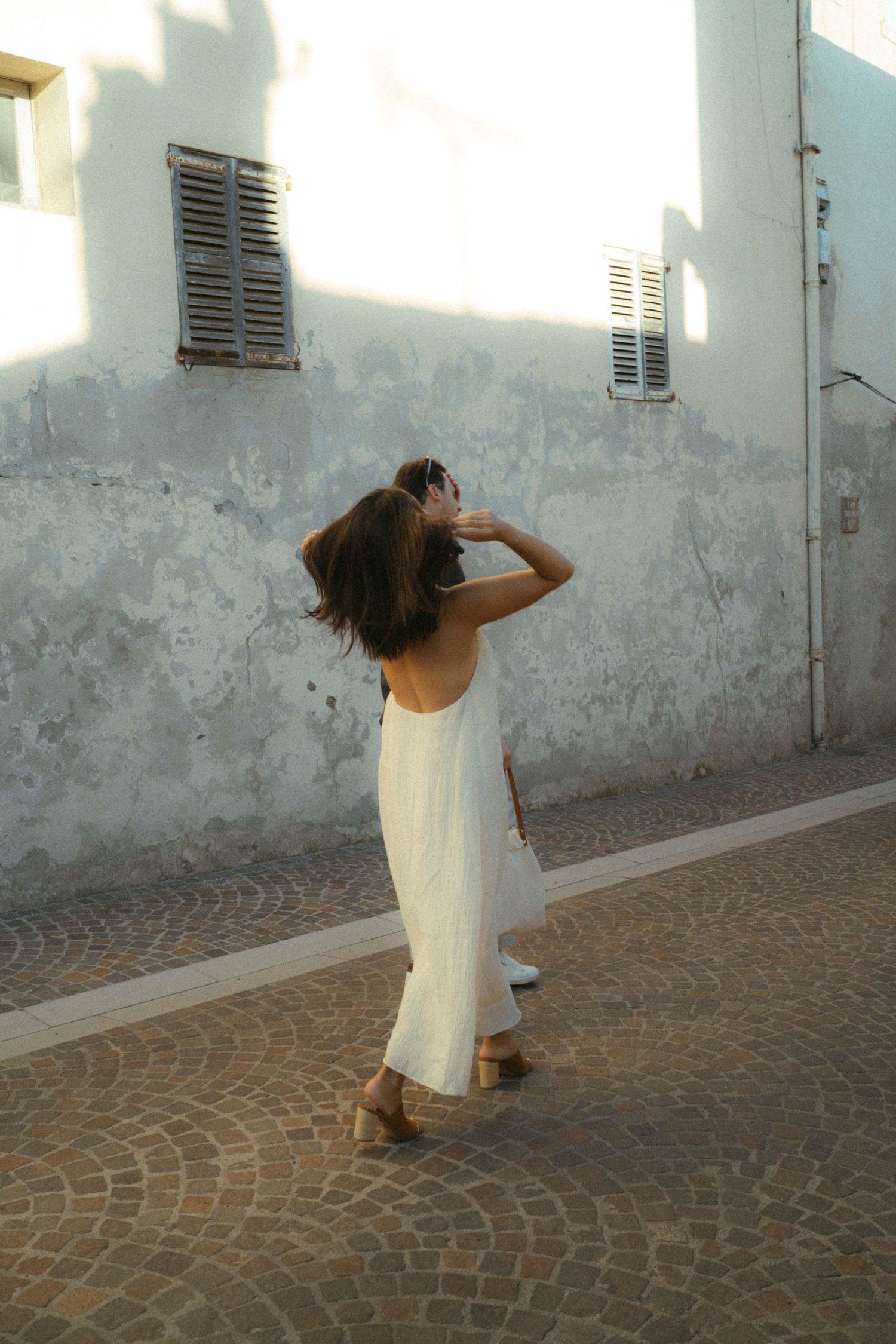 A sequence of images shows a person in a white dress walking along a cobblestone street next to a weathered wall.