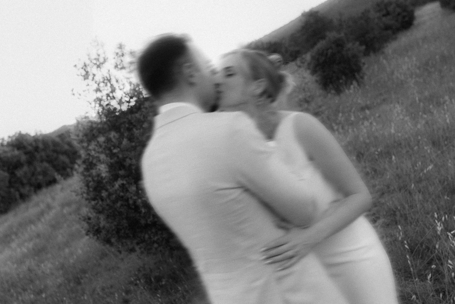 Black and white photo of a couple sharing a romantic kiss outdoors in a lush green field, dressed in white.