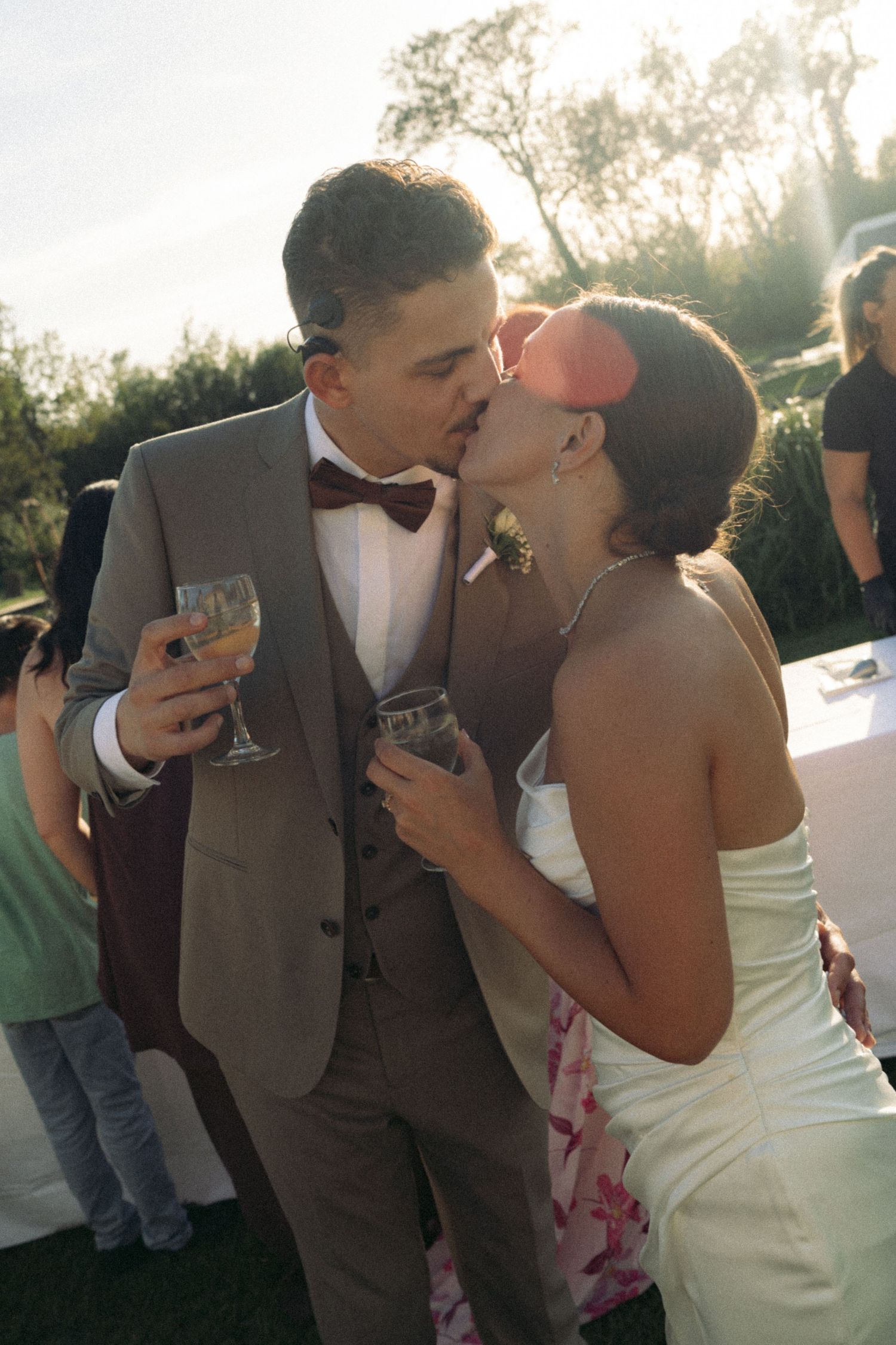 Newlywed couple sharing a kiss while holding wine glasses outdoors at their wedding reception.
