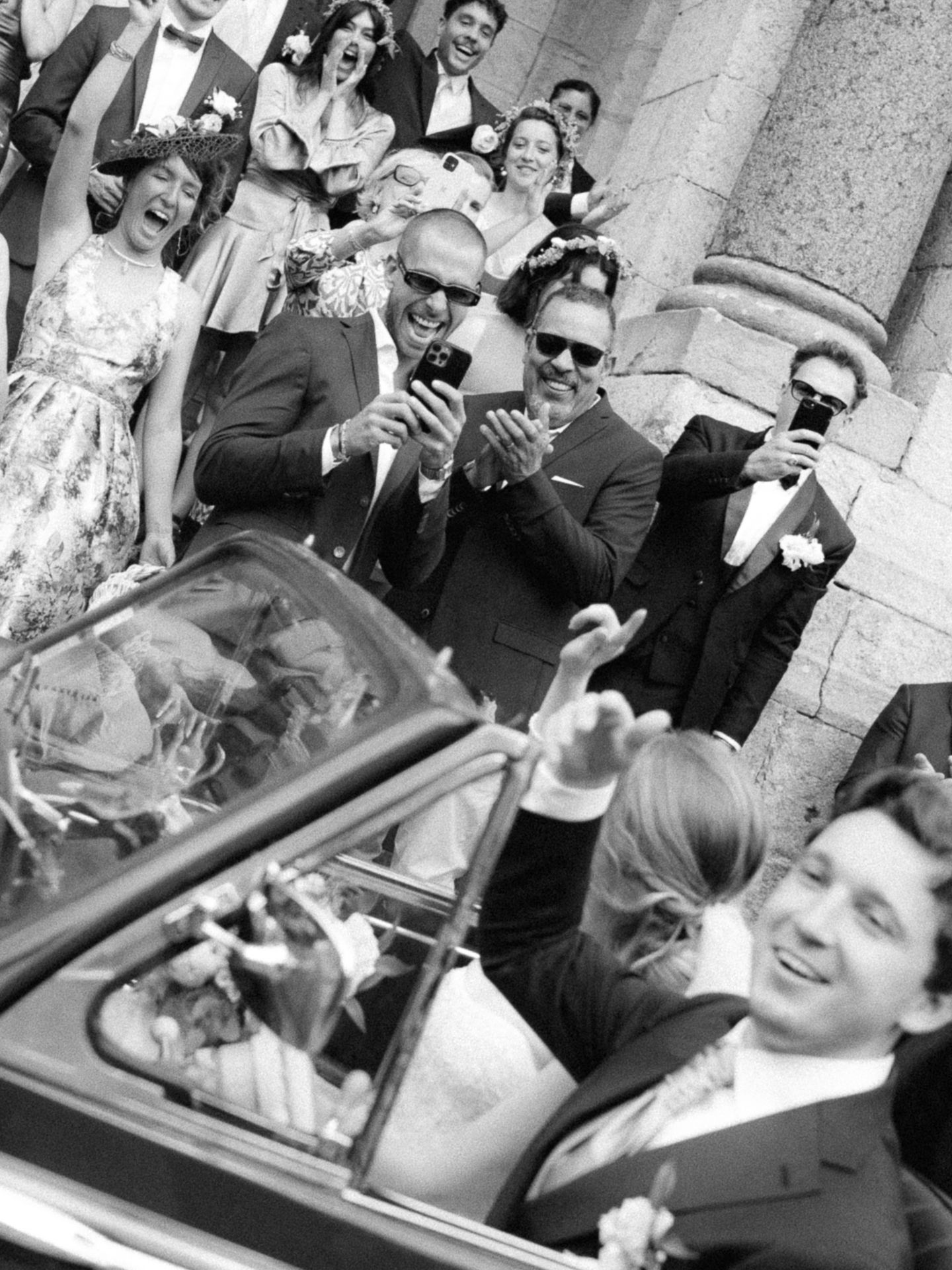 Black and white wedding photo of bride and groom in convertible car, waving to cheering guests outside church.
