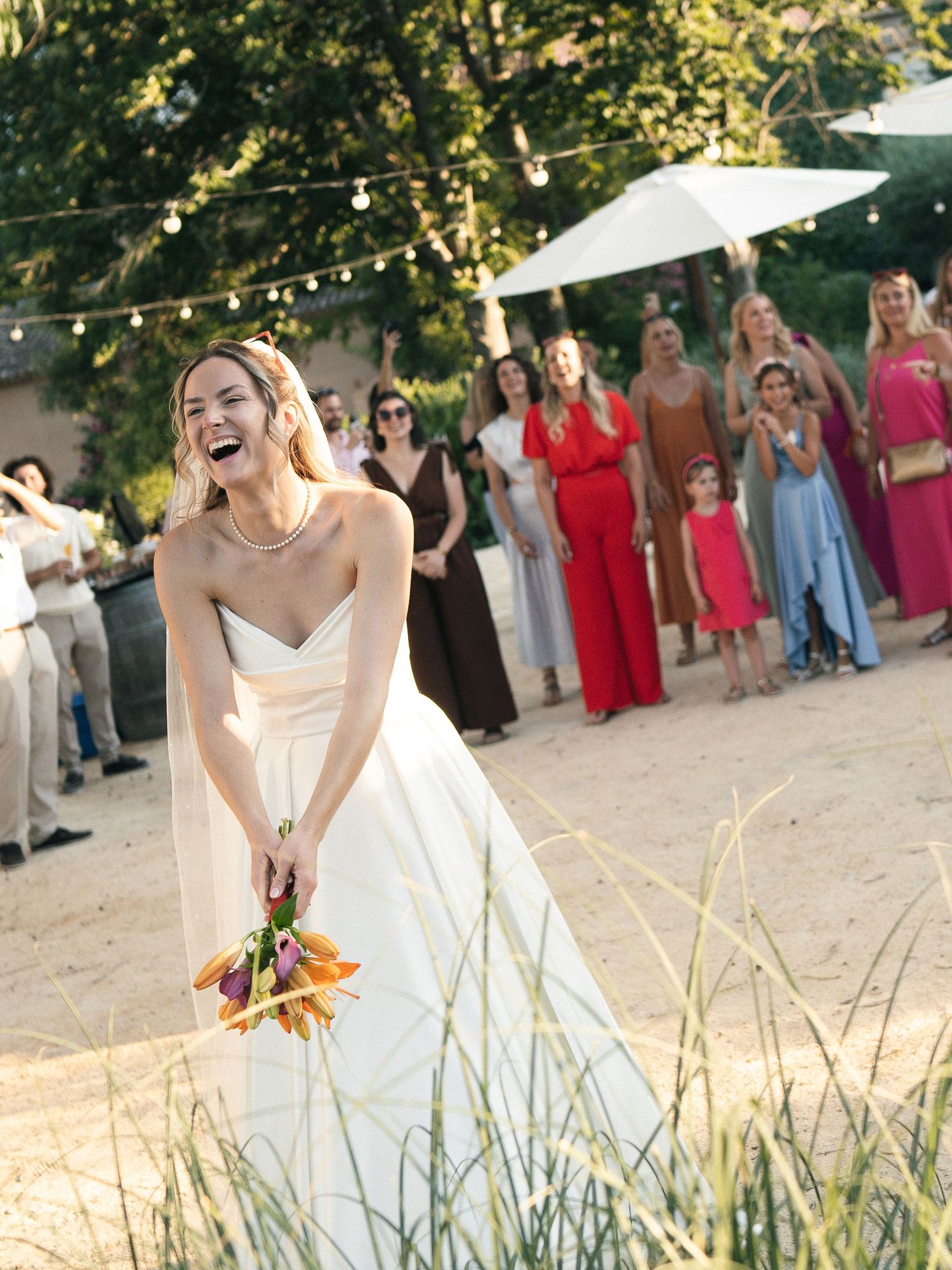 Laughing bride in white gown holds orange bouquet before tossing it to guests at outdoor wedding reception.